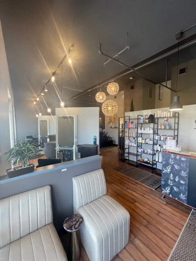 Interior of a modern salon or spa with beige upholstered chairs, wooden flooring, decorative lighting fixtures, and a display shelf with products.