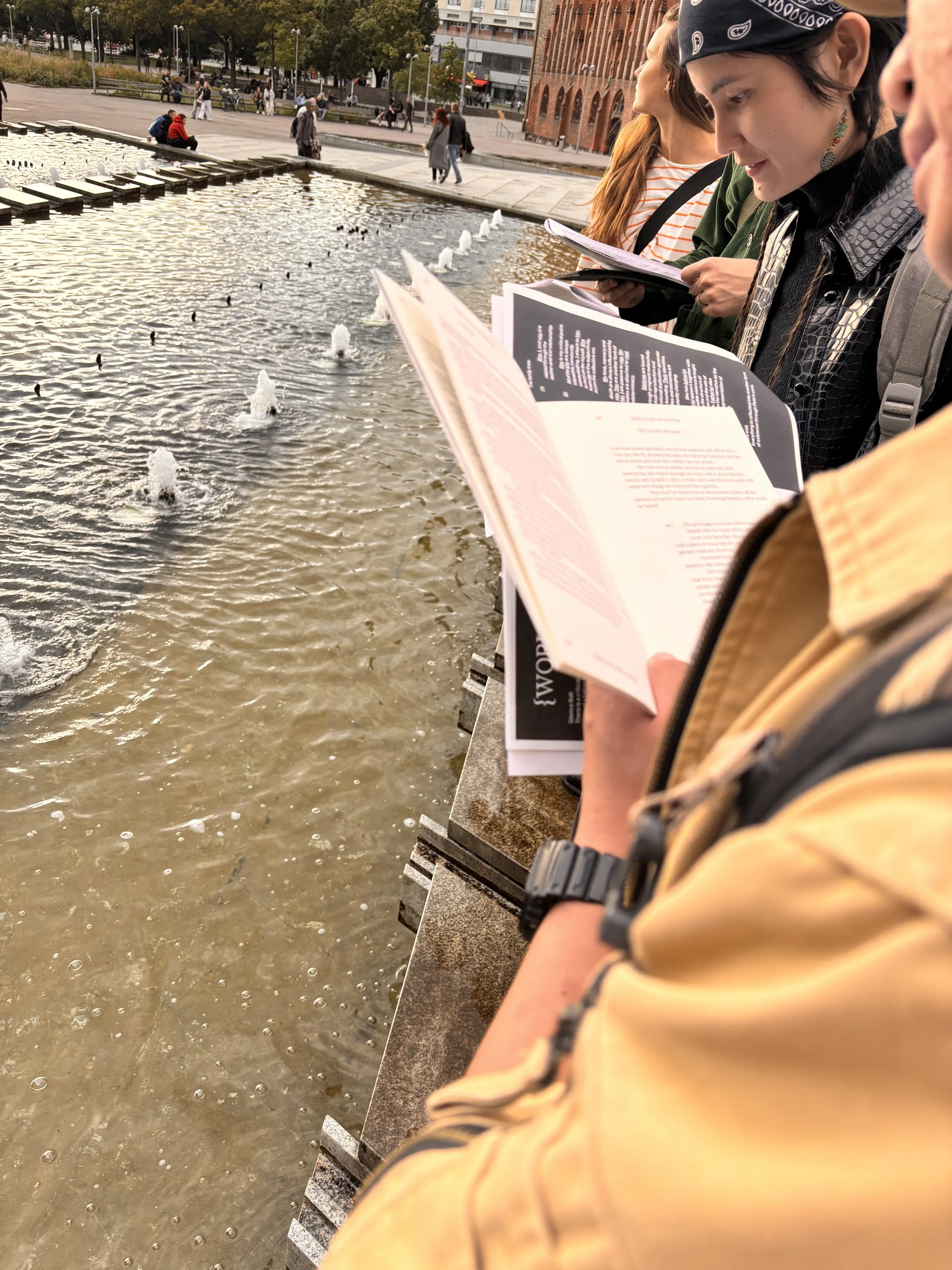 People standing along a water feature reading books near a fountain in an urban park.