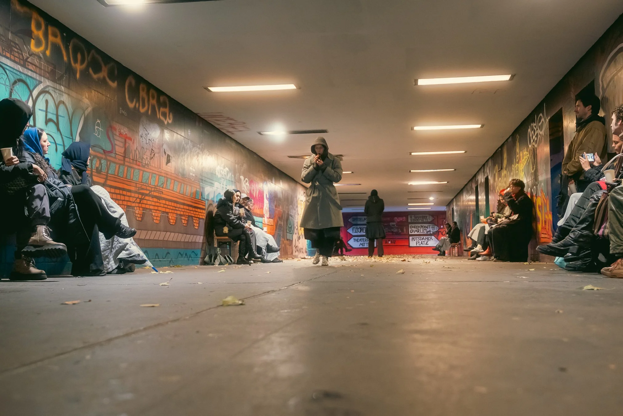 People sitting and standing along graffiti-covered walls in an underground tunnel, some taking photos, others waiting or walking.