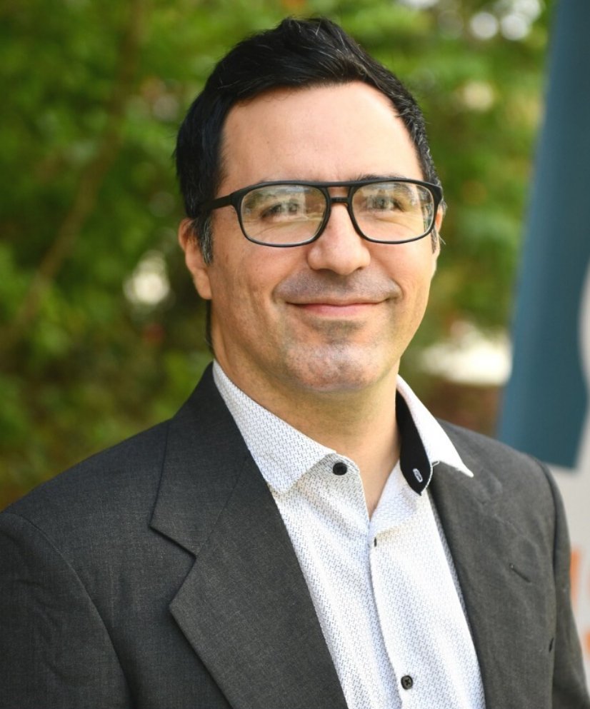 A smiling man with black hair, glasses, and a small beard, wearing a dark blazer and a white shirt, standing outdoors with blurred green foliage in the background.
