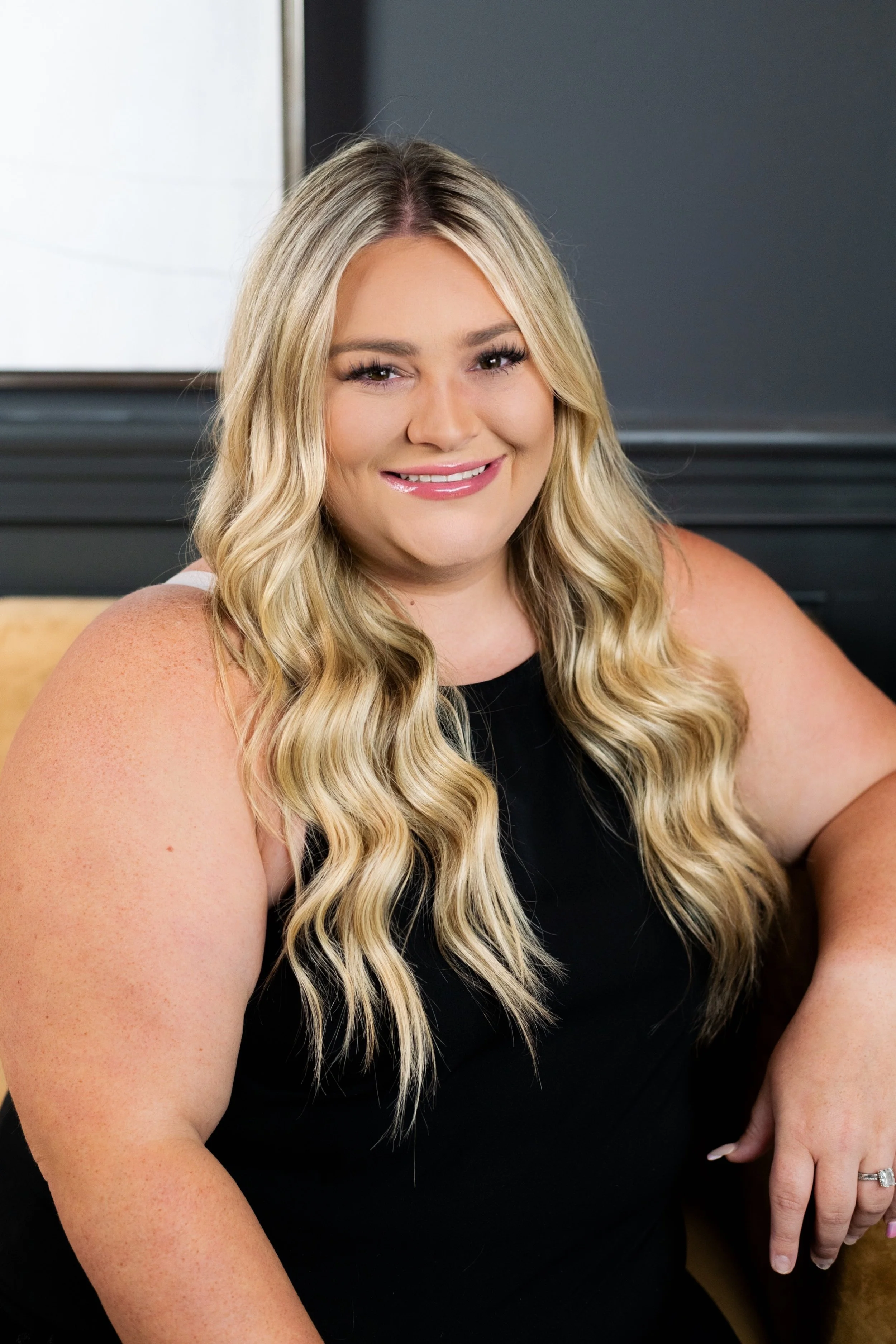 A woman with long blonde wavy hair, wearing a sleeveless black top, smiling and seated indoors.