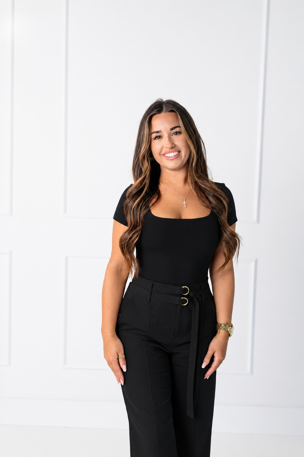 Young woman with long, wavy brown hair sitting on a beige couch near a window with black trim, wearing a black sleeveless top and a necklace, smiling softly.
