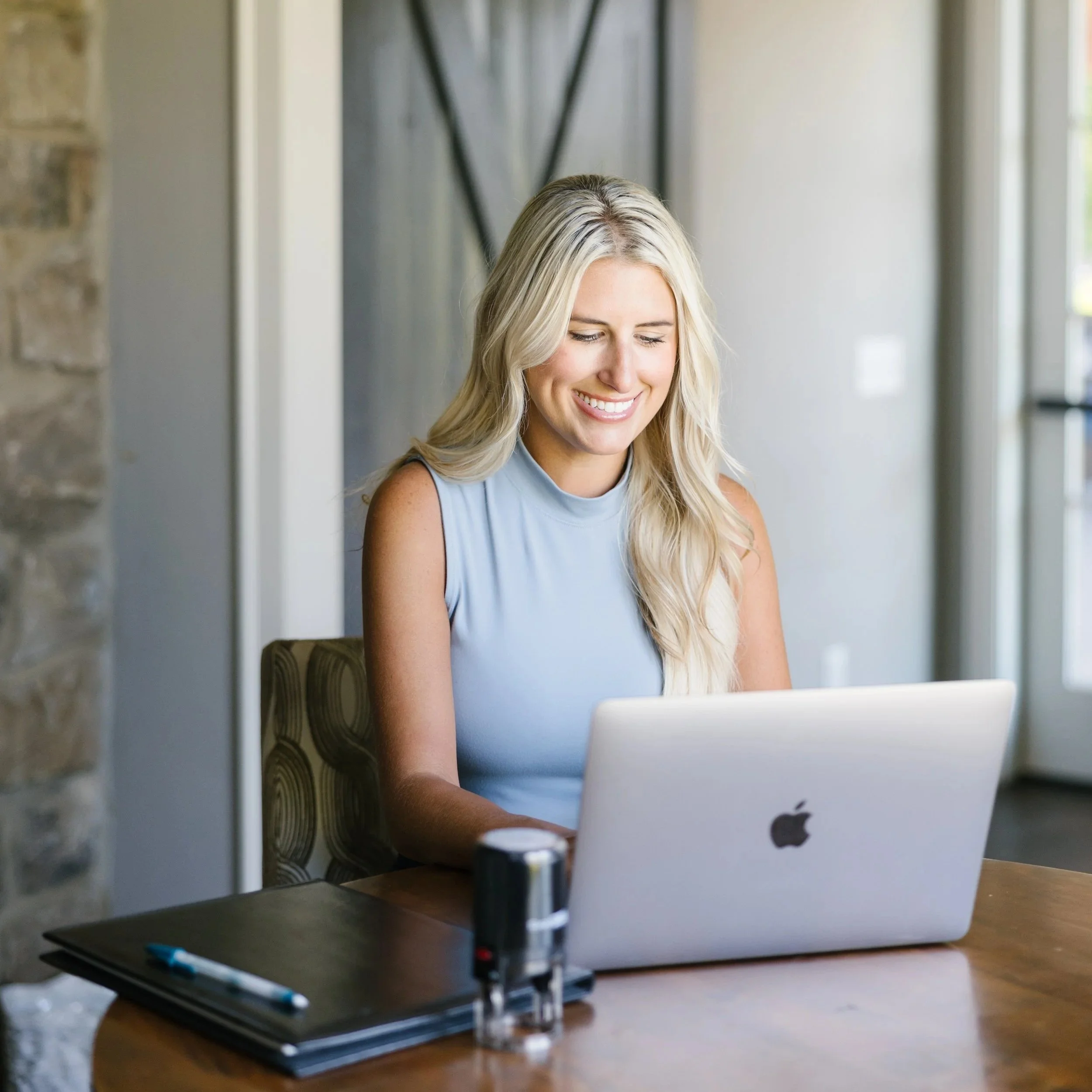 A woman with blonde hair smiling while working on a laptop at a desk. There are pens and a notebook on the desk, and a microscope in front of the laptop. The background shows a window and a wall.