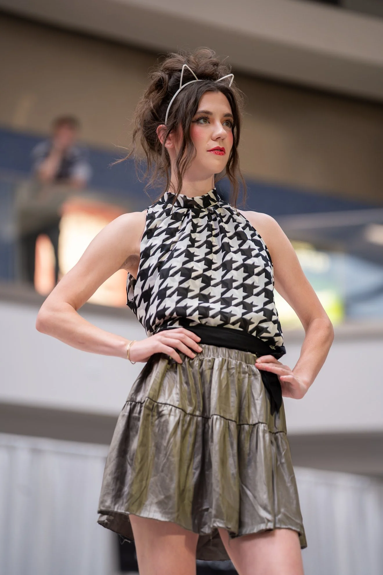 A woman with dark hair styled in messy updo, wearing a cat ear headband, a black and white houndstooth sleeveless top, and a metallic gold and black pleated skirt, standing with her hands on her hips in an indoor setting.