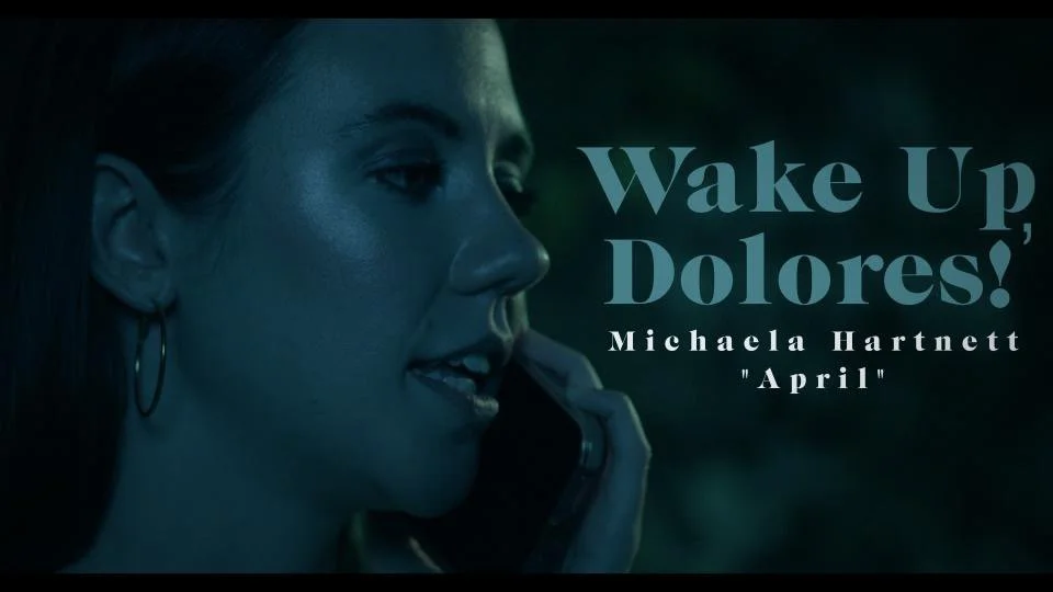 A woman with hoop earrings talking on a cellphone at night, with the text "Wake Up, Dolores! Michaela Hartnett 'April'" displayed on the right side.