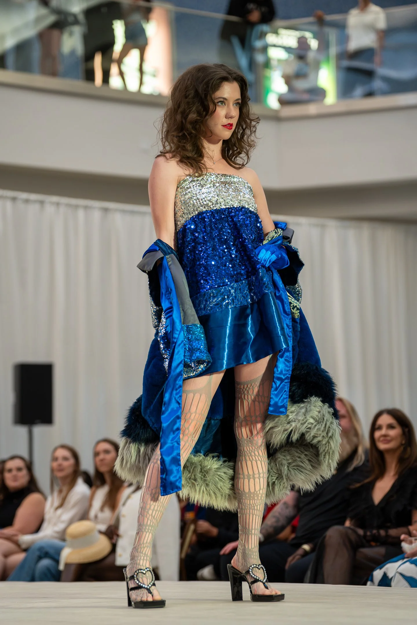 A fashion runway show featuring a model in a sparkling, strapless dress with blue, gold, and silver sequins, paired with fishnet tights, furry green leg warmers, and black high-heeled sandals with heart-shaped embellishments. The audience is visible 