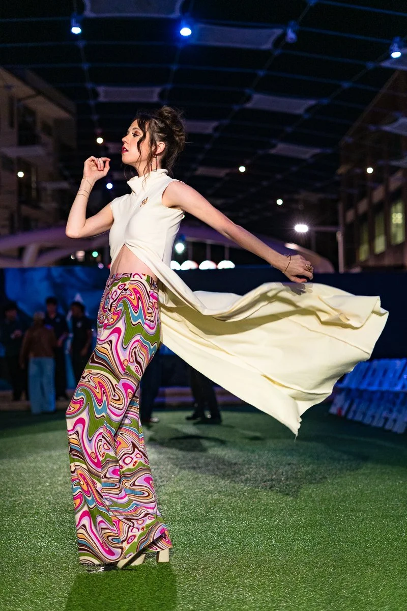 A woman in colorful, patterned wide-leg pants and a sleeveless top dancing at a nighttime event with a dark canopy overhead and people in the background.