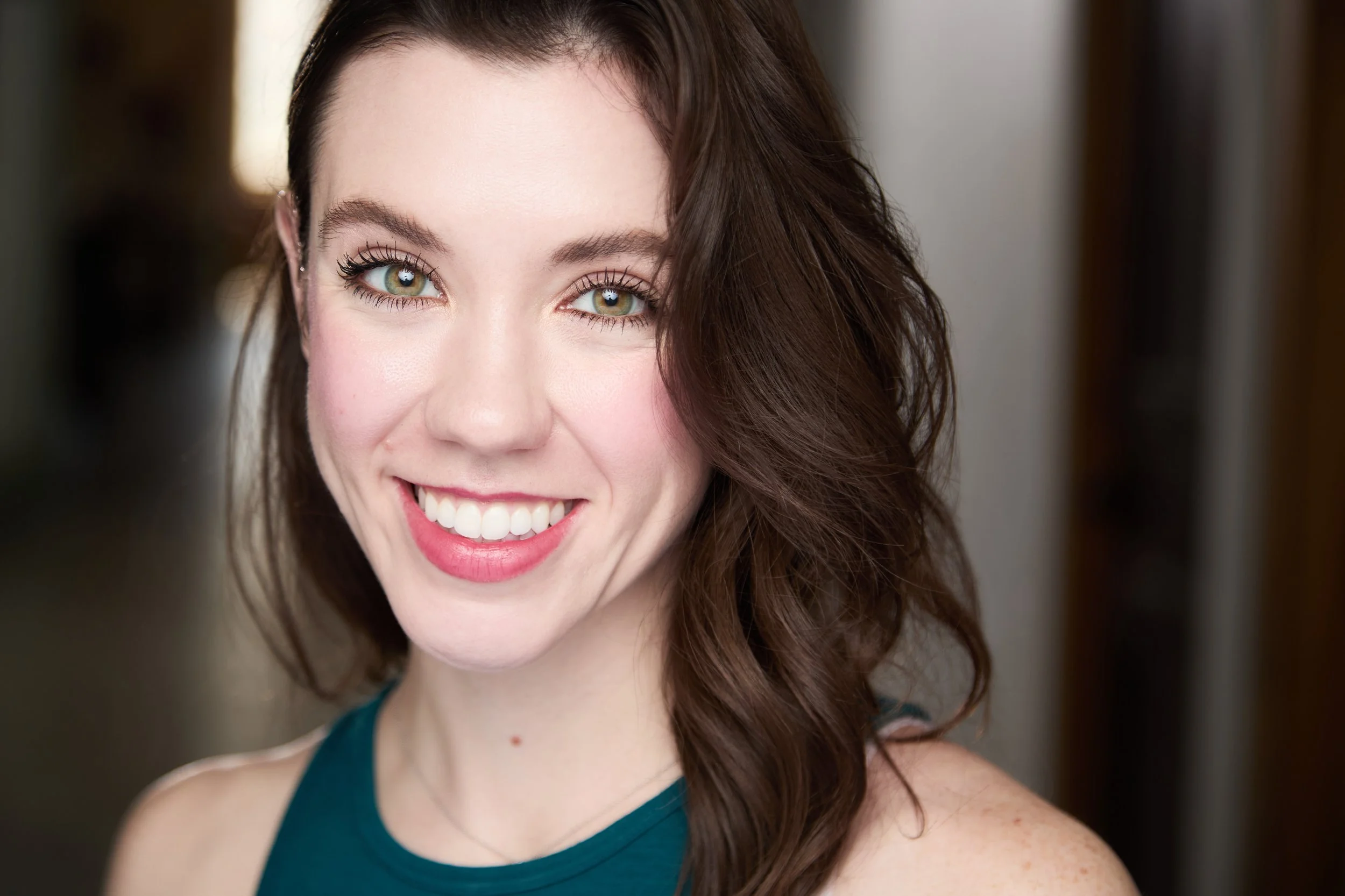 Close-up of a smiling woman with curly brown hair, blue eyes, and light skin, wearing a sleeveless teal top.