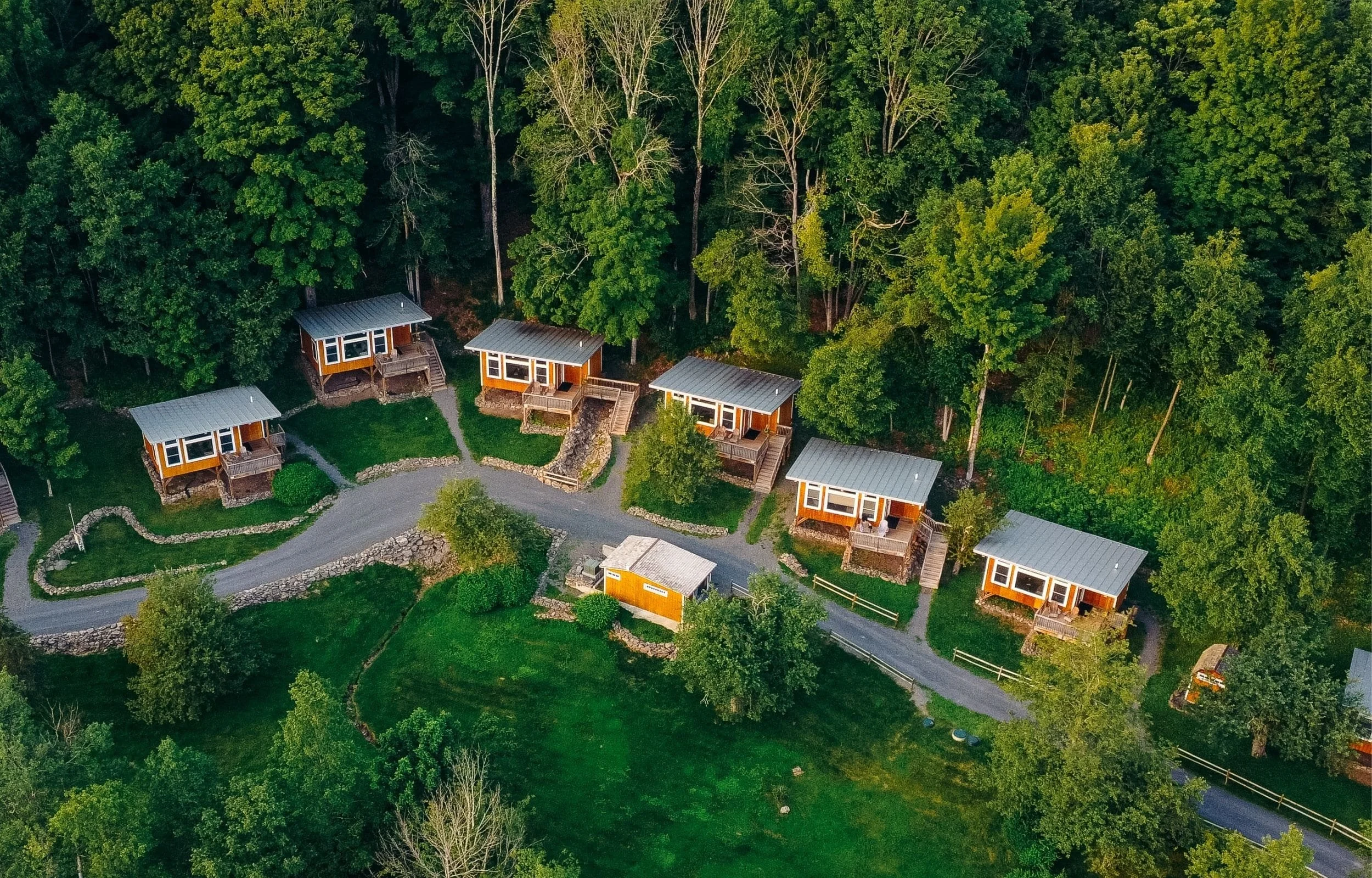 Aerial view of several small wooden cabins with metal roofs lined along a winding road in a green, wooded area.