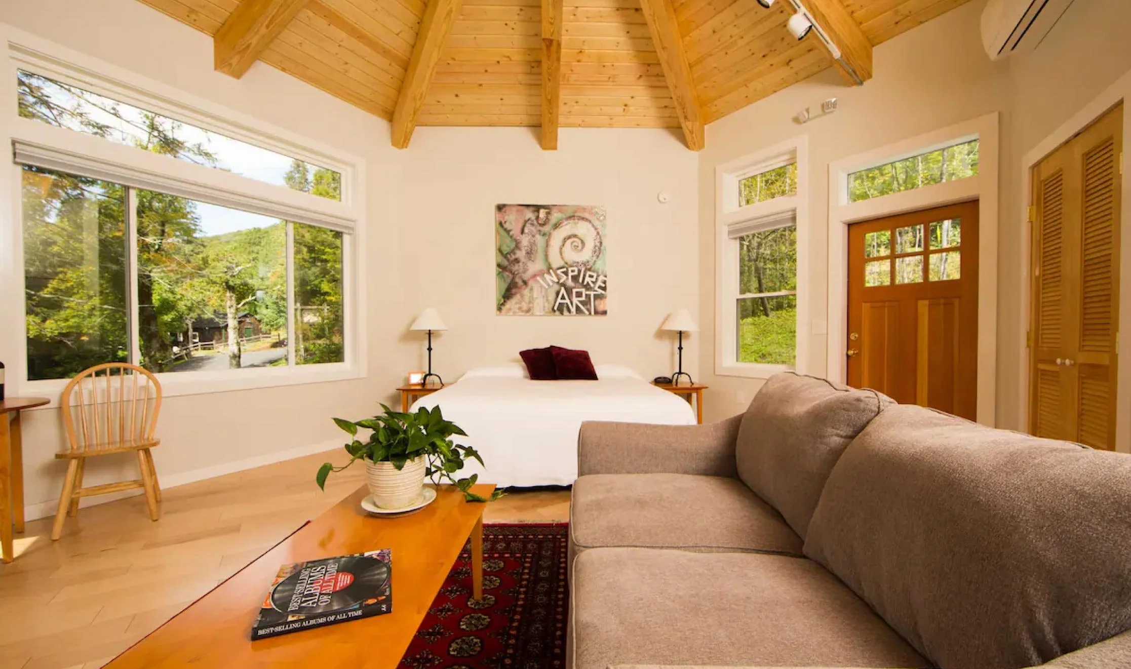 Living room with large windows, a bed with two lamps, a wooden door, a beige sofa, a coffee table with a plant and a magazine, and a colorful artwork that says 'Inspire Art' on a wood ceiling.