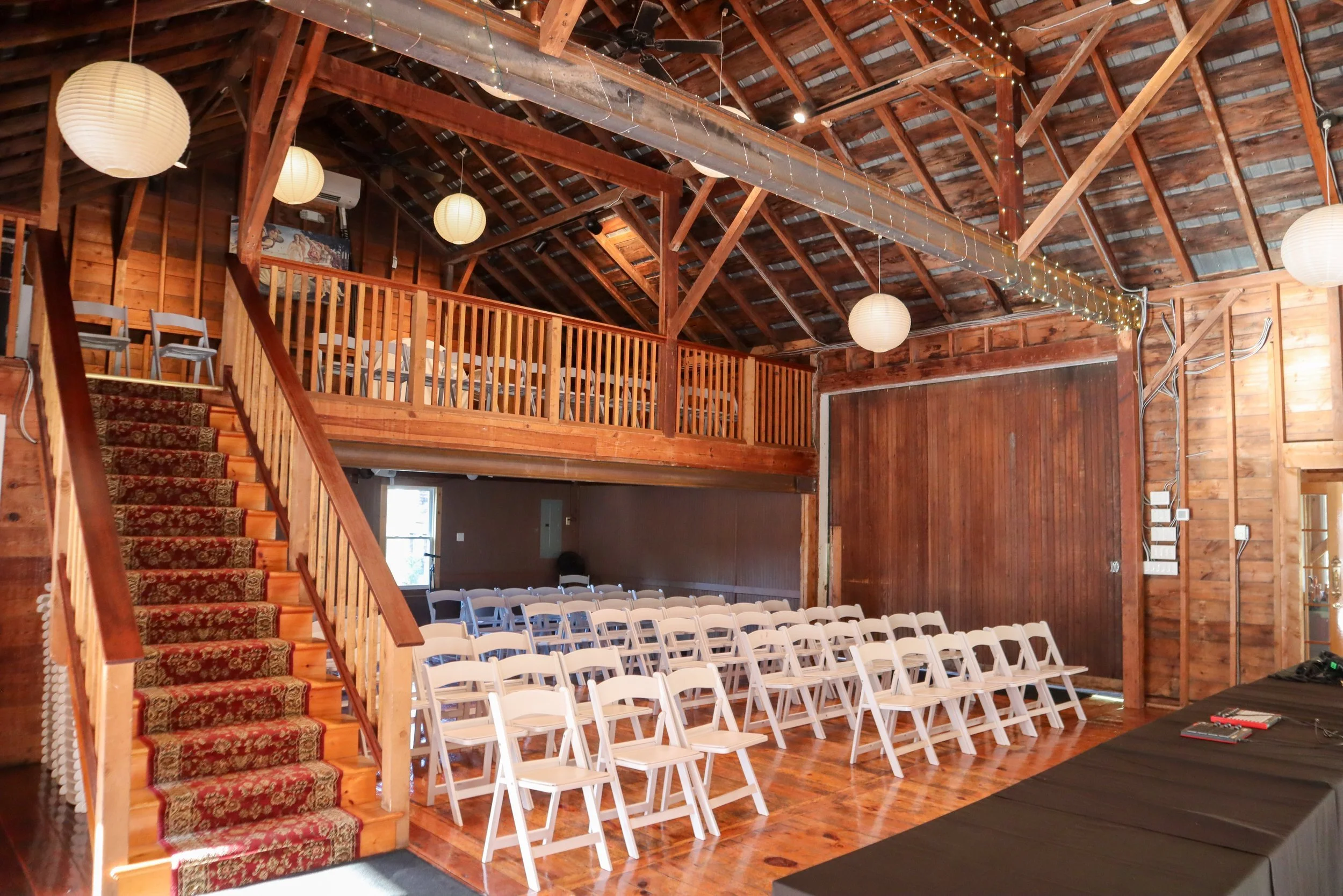 Interior of a rustic event space with wooden walls and ceiling, white chairs arranged in rows facing a stage area, a staircase with patterned carpet on the left, hanging white paper lanterns, string lights, and a balcony area with additional chairs.