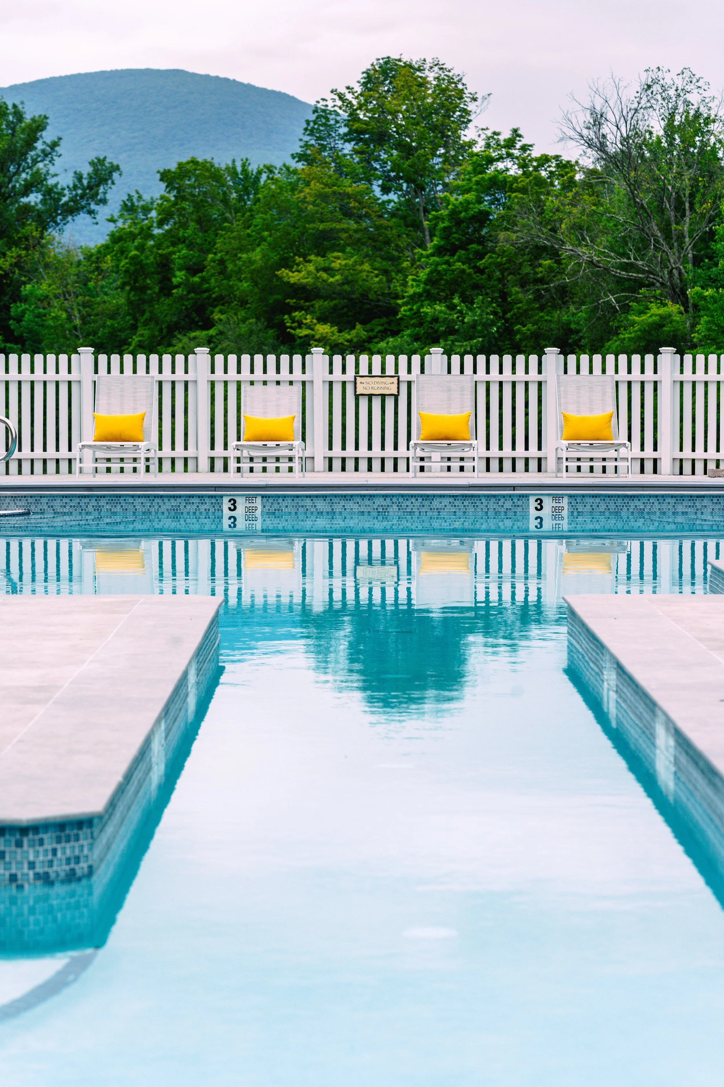 A pool area with a white fence and four white chairs with yellow pillows, green trees, and mountains in the background.