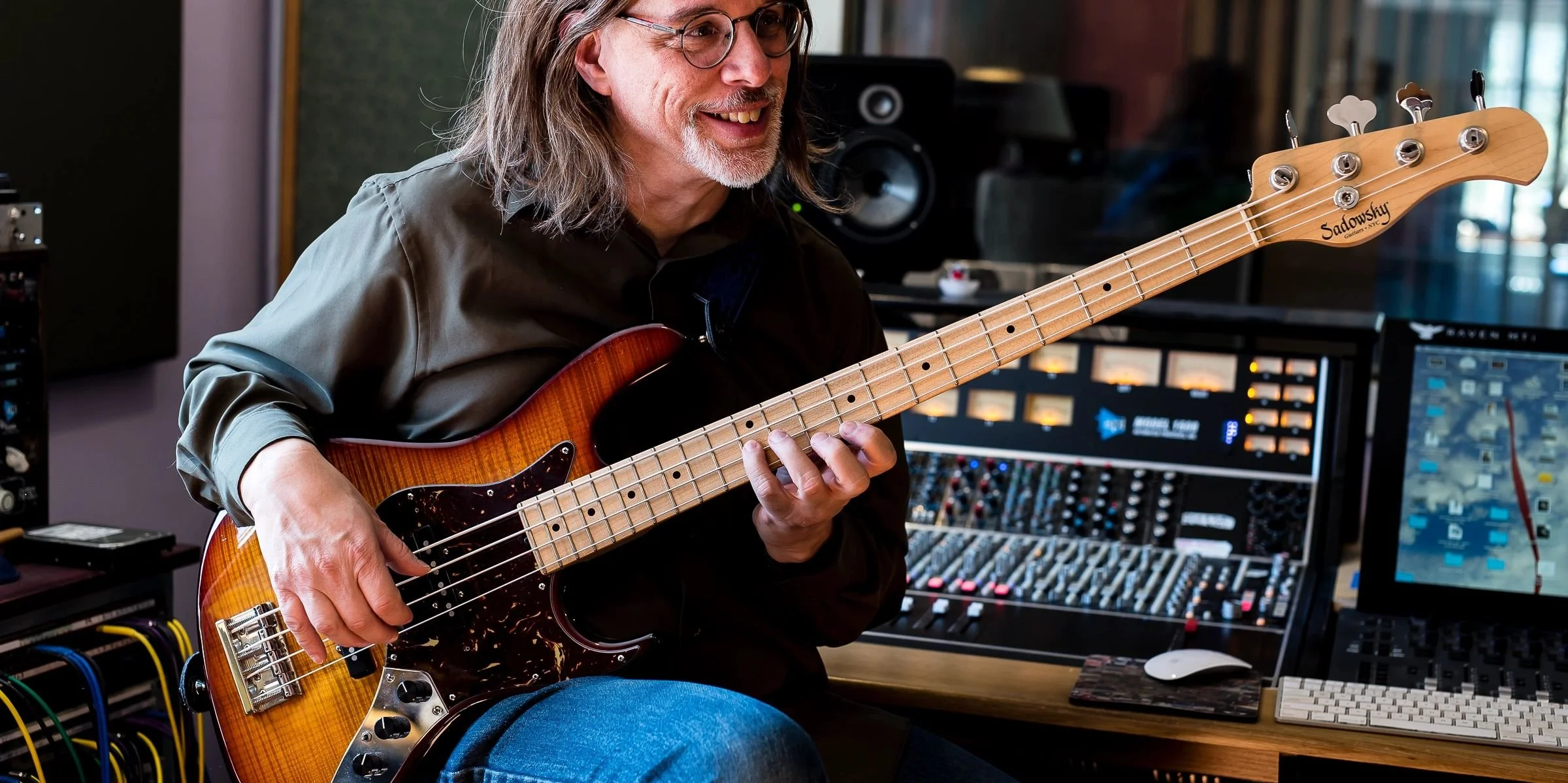 A man with long gray hair and glasses playing an electric bass guitar in a professional music studio.