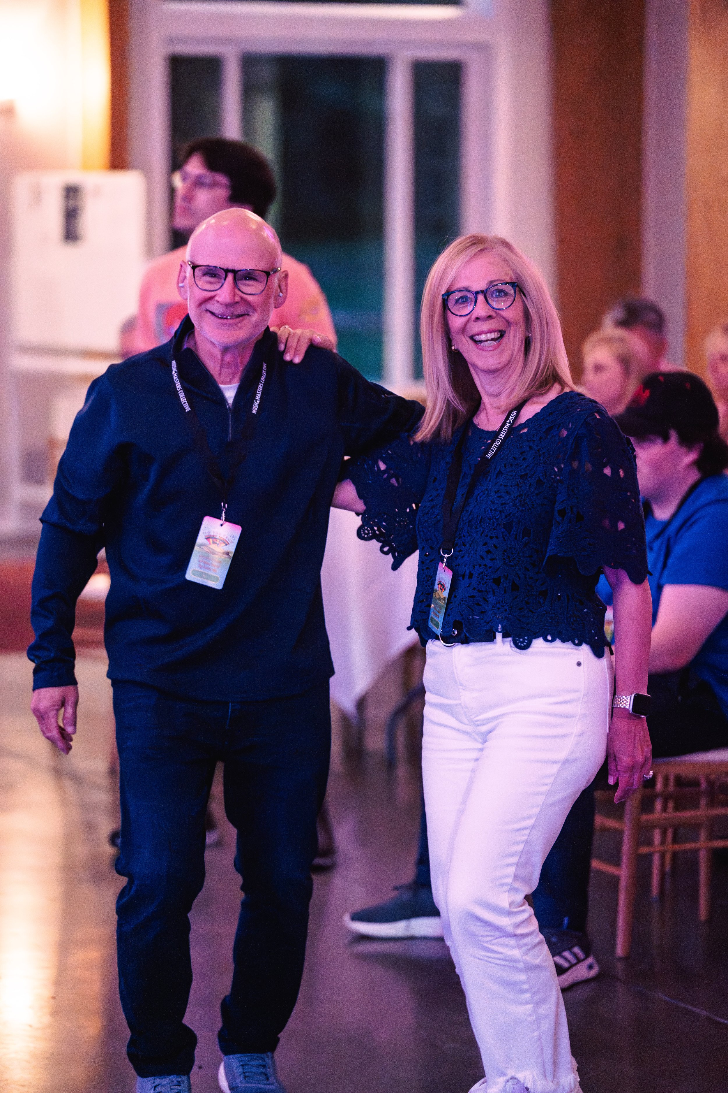 Two smiling adults, a man and a woman, standing together with their arms around each other at an indoor event, with other people in the background.