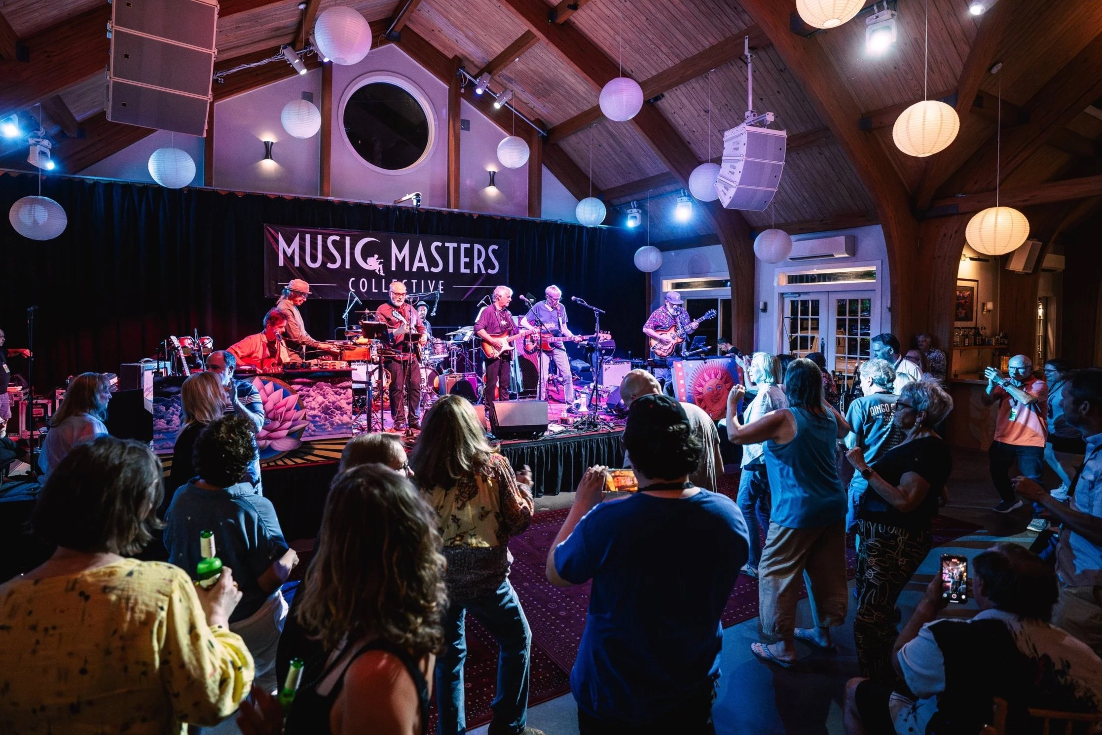 Audience dancing and enjoying live music performance at an indoor venue with a band on stage and a 'Music Masters Collective' banner.