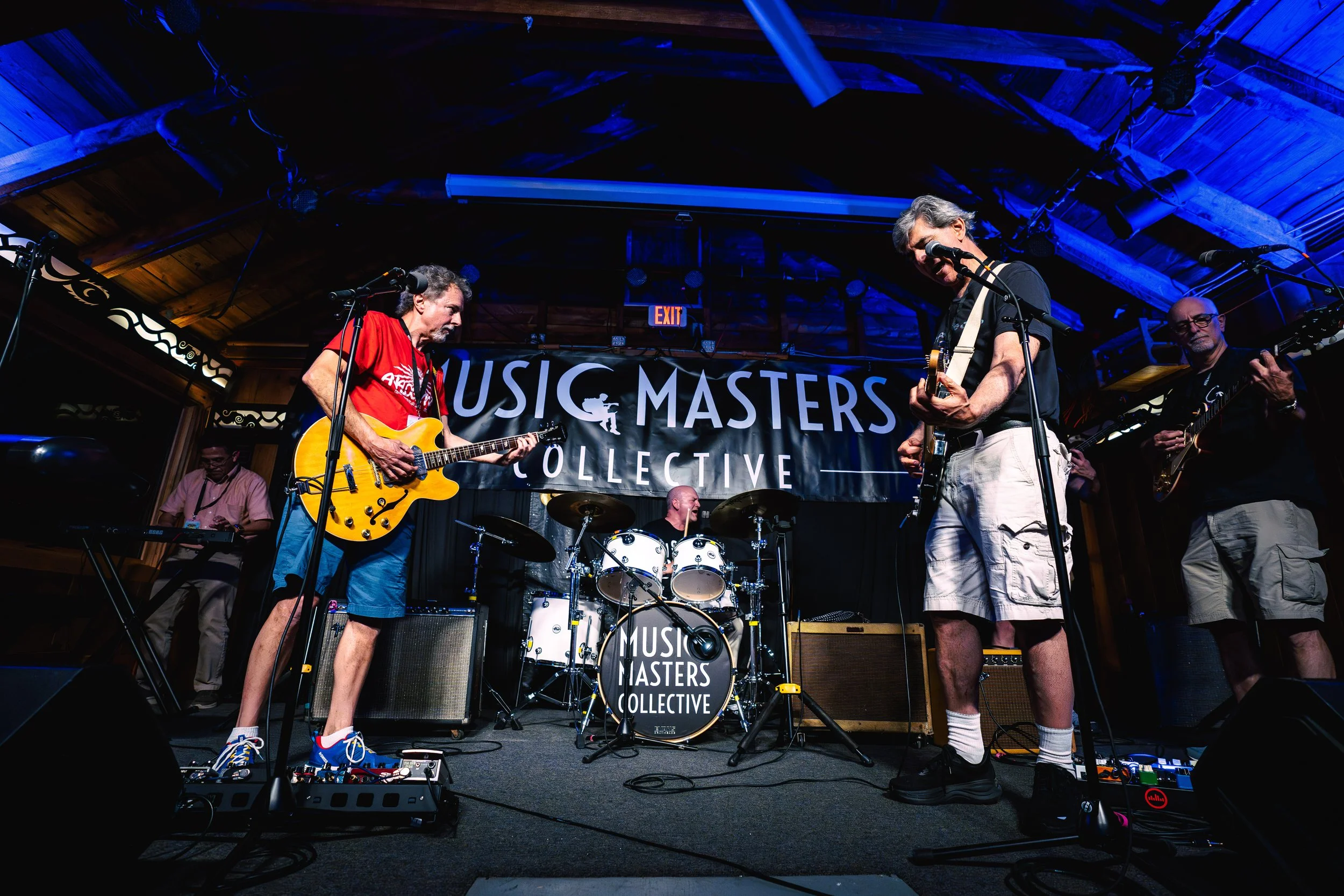 A band performing live on stage with a banner behind them that reads 'Music Masters Collective'. The band includes a drummer, two guitarists, and a keyboardist, with the guitarists in the foreground and the keyboardist in the background. The stage is indoors with wooden walls and ceiling, illuminated by stage lights.