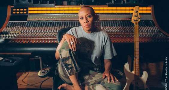 A woman sitting in a music studio, surrounded by a large mixing console and a guitar.