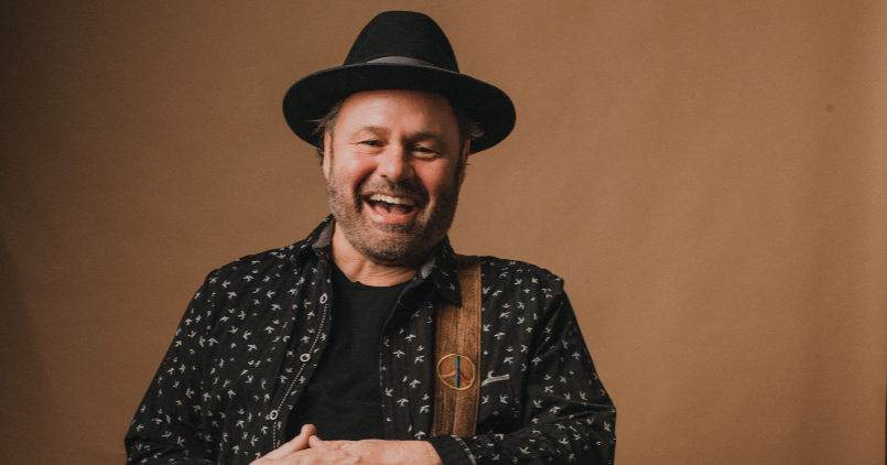 A man with a beard and mustache wearing a black hat, black patterned shirt, and brown suspenders, smiling and laughing against a plain brown background.
