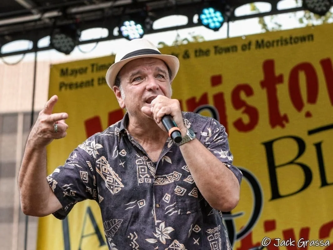 A man wearing a Hawaiian shirt and a fedora hat holds a microphone and speaks on stage at an outdoor event with a yellow banner in the background.