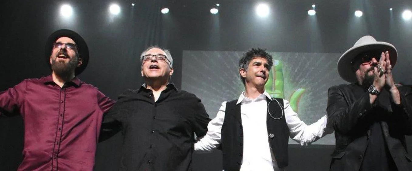 Four men standing on stage, holding hands and singing or speaking into microphones, with a large screen in the background displaying a green banana icon. The men are dressed in casual and semi-formal attire, with hats and glasses.