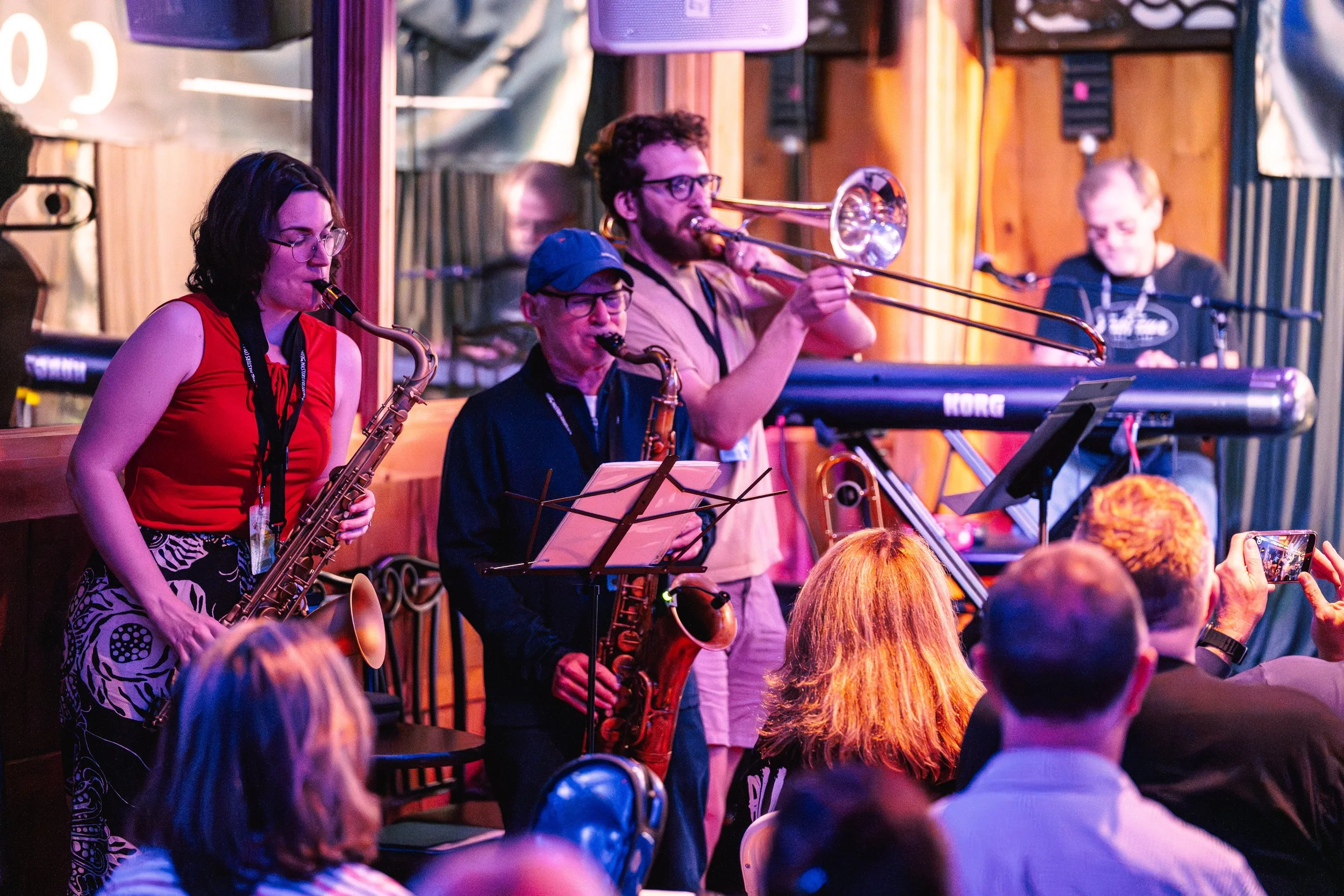 A jazz band performing on stage with a saxophone, a trombone, a keyboard, and a microphone, while an audience is watching and recording the performance.