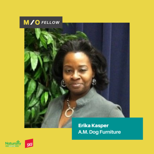 A woman with dark hair wearing a gray blazer and jewelry standing in front of green plants and a blue curtain, with a yellow border and text indicating her name as Erika Kasper and her company, A.M. Dog Furniture.