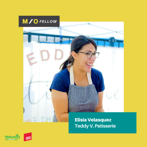 A woman with glasses and dark hair tied back, wearing a blue shirt and gray apron, smiling at a Teddy V. Patisserie booth at a MOFELLOW event.