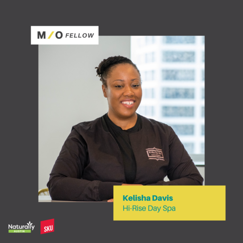 Portrait of Kelisha Davis, Hi-Rise Day Spa, smiling with dark hair in a bun, wearing a black spa uniform, inside a modern office with windows showing city buildings.
