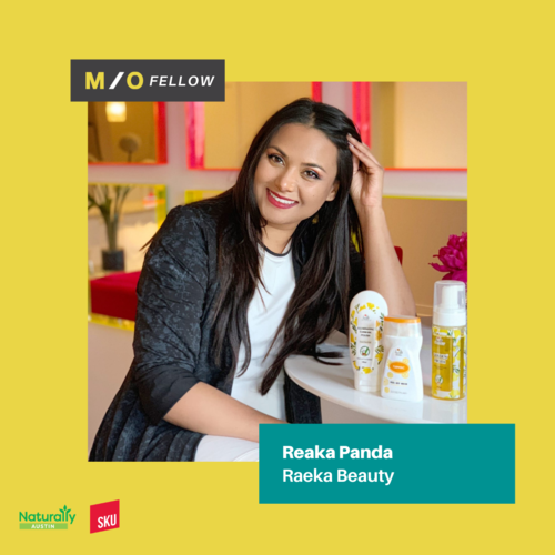 Smiling woman with long dark hair sitting at a table with skincare products in front of her, indoors with yellow walls and pink accents.