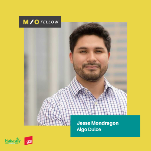 A professionally dressed man with dark hair and a beard smiling outdoors against a cityscape background, side profile, with a yellow border and text overlay.