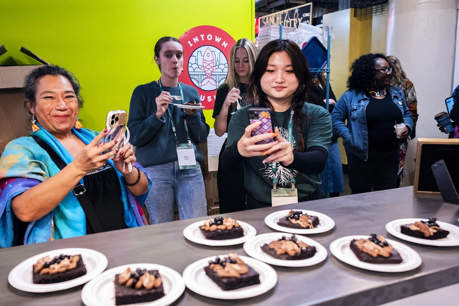 Group of women at a food event, taking photos of desserts on a table, with a green wall and logo in the background.