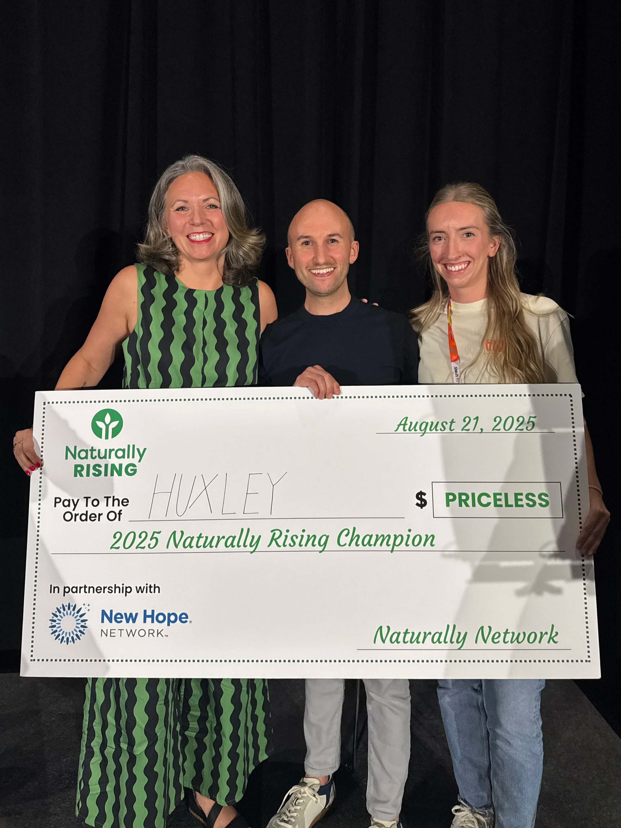 Three smiling people holding a large check, standing in front of a black backdrop. The check is dated August 21, 2025, and is for the title of 2025 Naturally Rising Champion, associated with Naturally Network and New Hope Network.