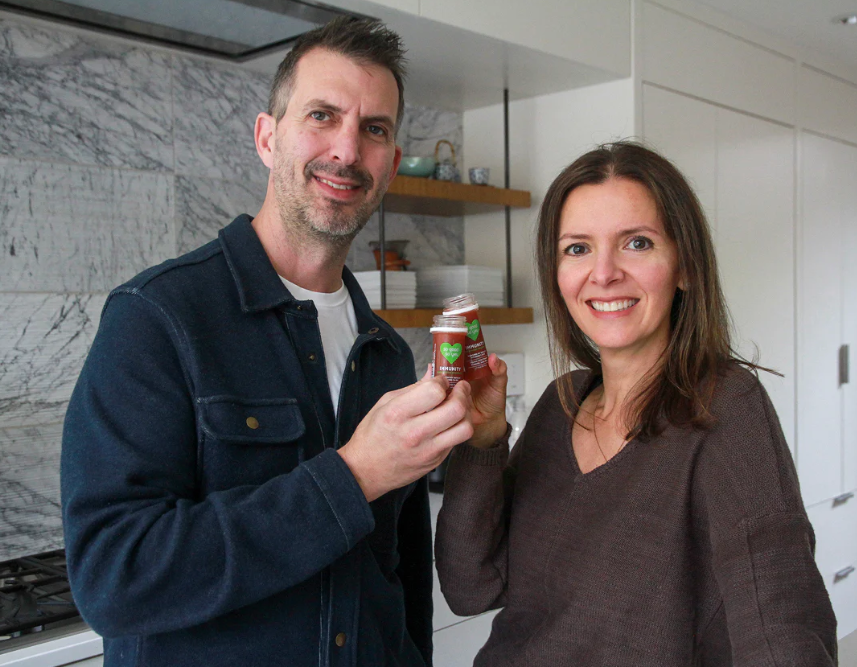 A smiling man and woman holding a jar of strawberry jam in a modern kitchen.