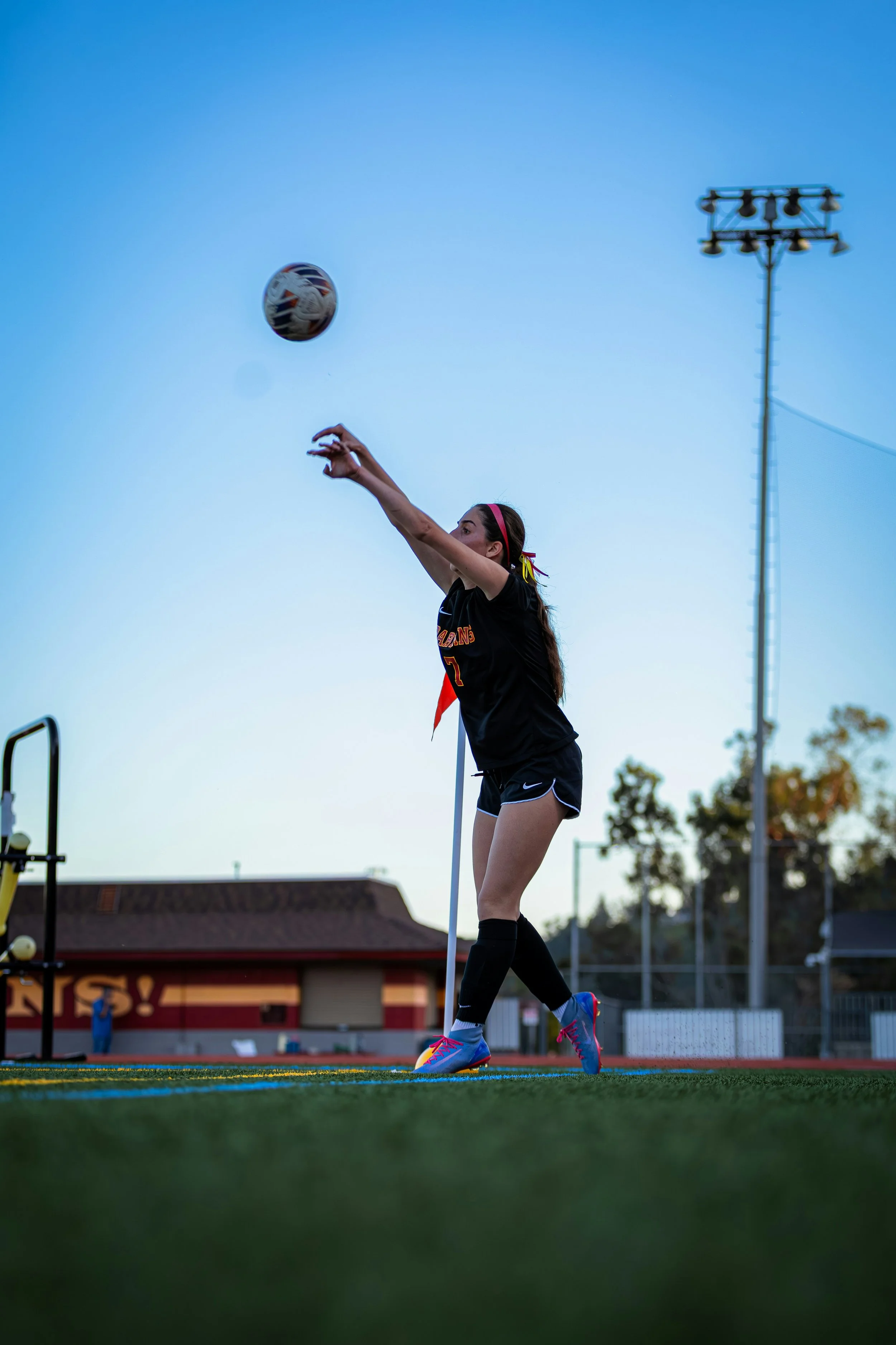 A female soccer player in black uniform hitting a soccer ball on a field during dusk.