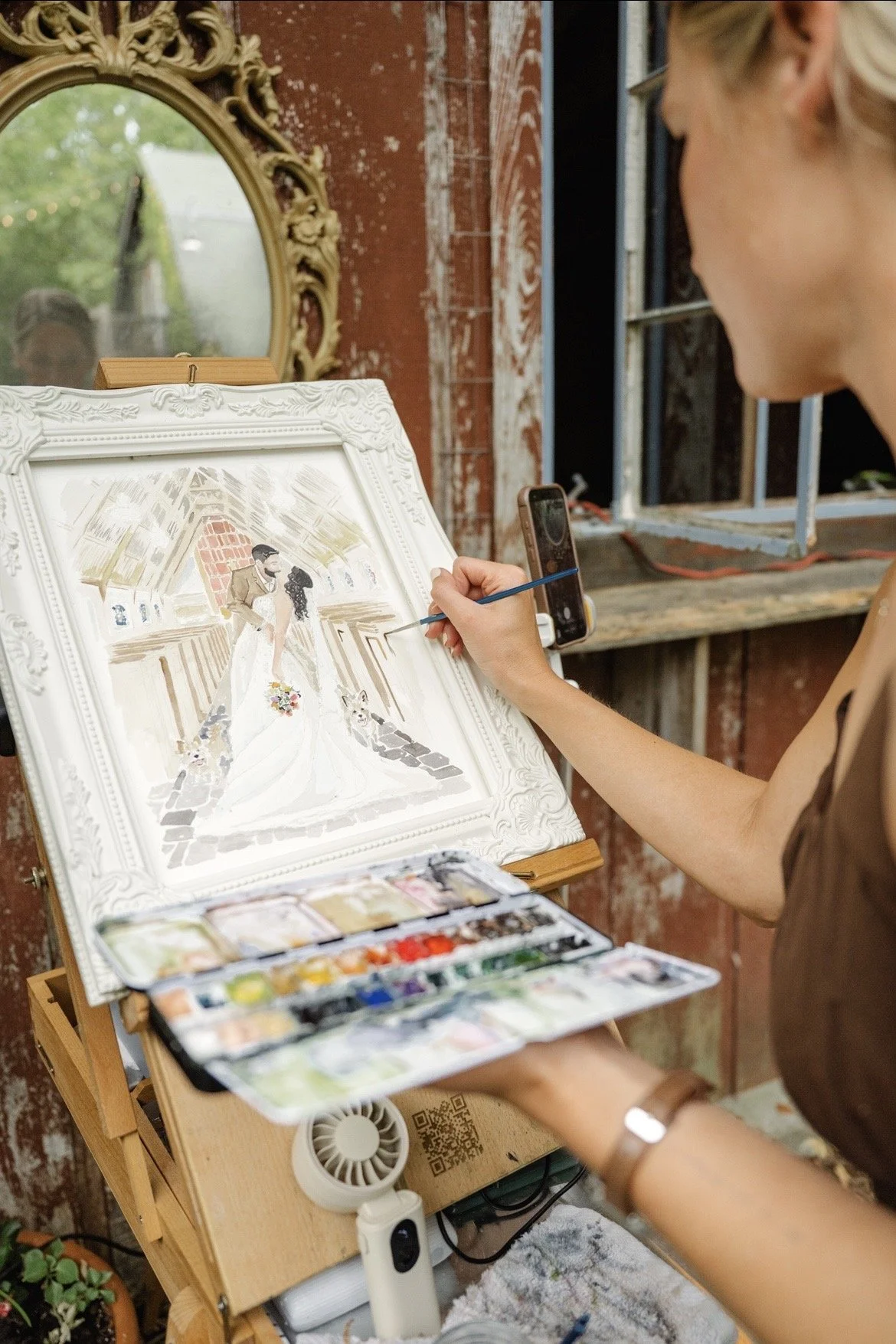 An artist painting a wedding scene on a canvas, showing a couple in wedding attire with a dog, inside a rustic building with exposed brick and a decorative mirror on the wall.
