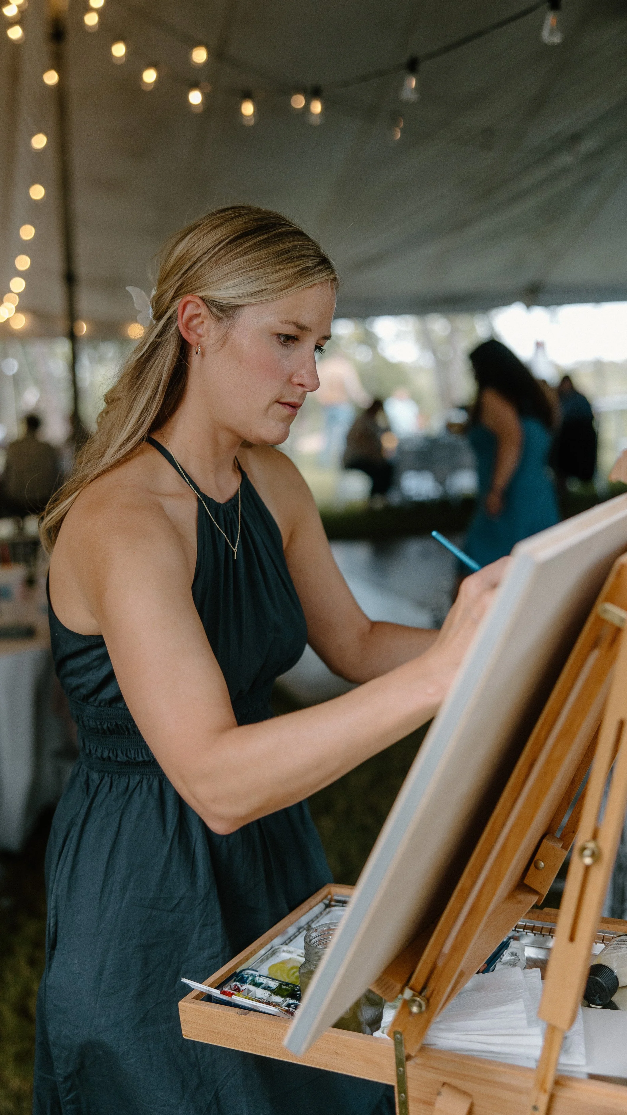 A woman in a dark blue dress live painting on a canvas at a wedding event under a tent with string lights and other people in the background.