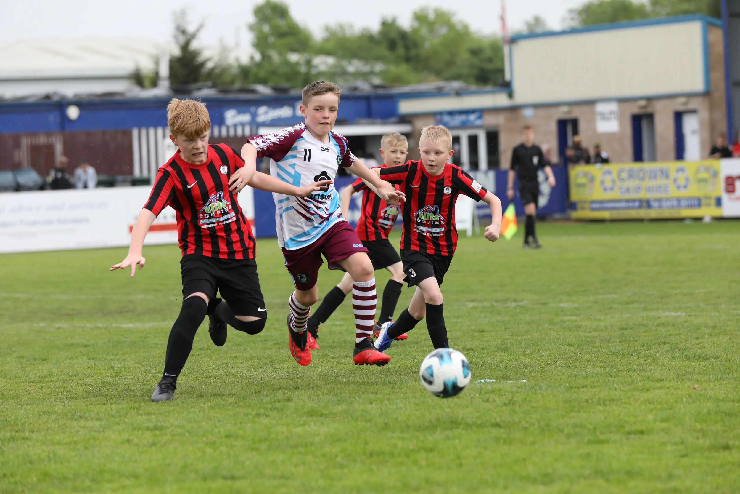Four young boys playing soccer on a grass field. Two are wearing red and black striped jerseys, and one is in a white and maroon jersey. They are actively chasing the ball during a game.