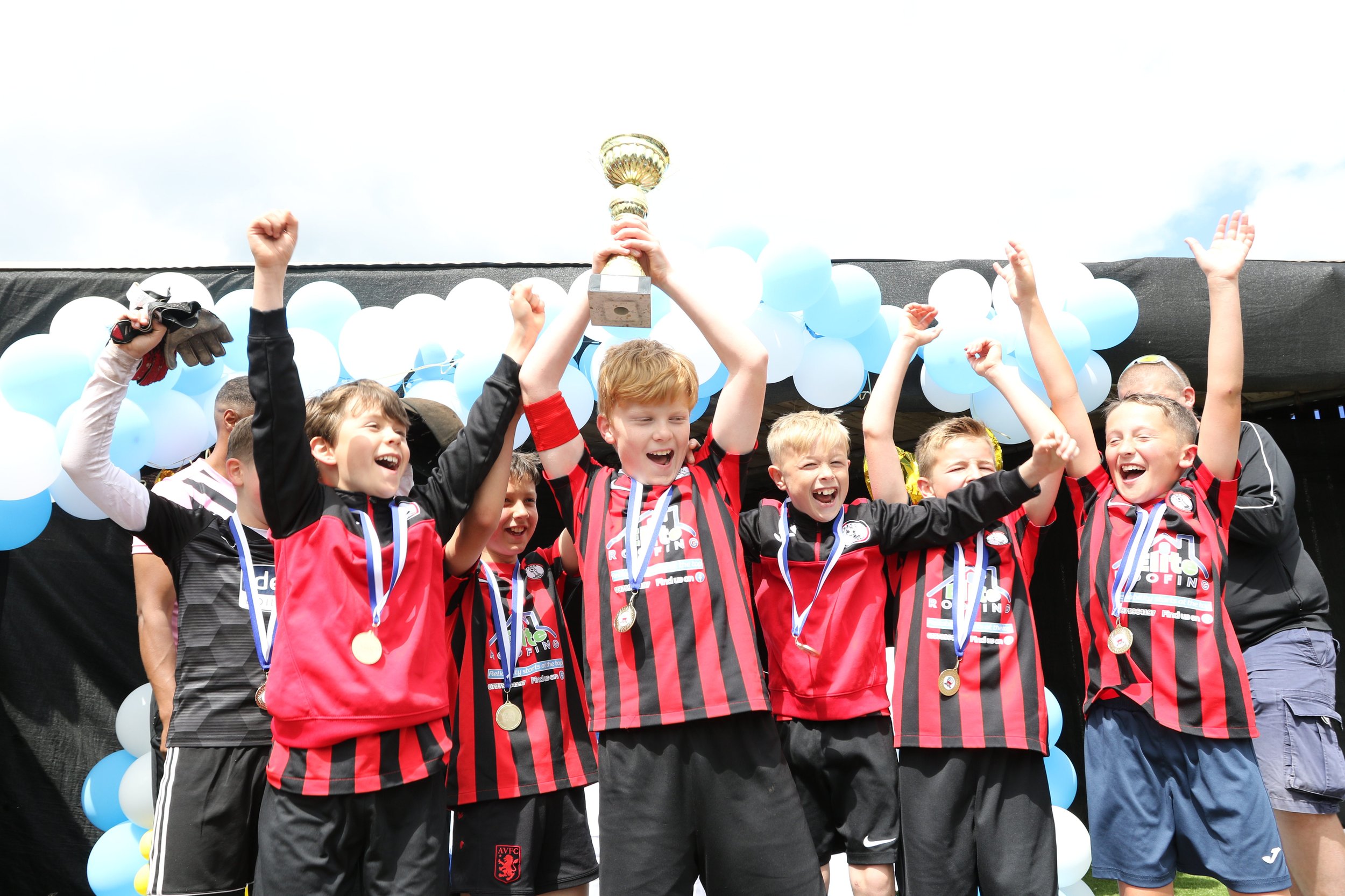 Kids celebrating on a soccer field with a trophy, medals, and team uniforms, under blue and white balloons.