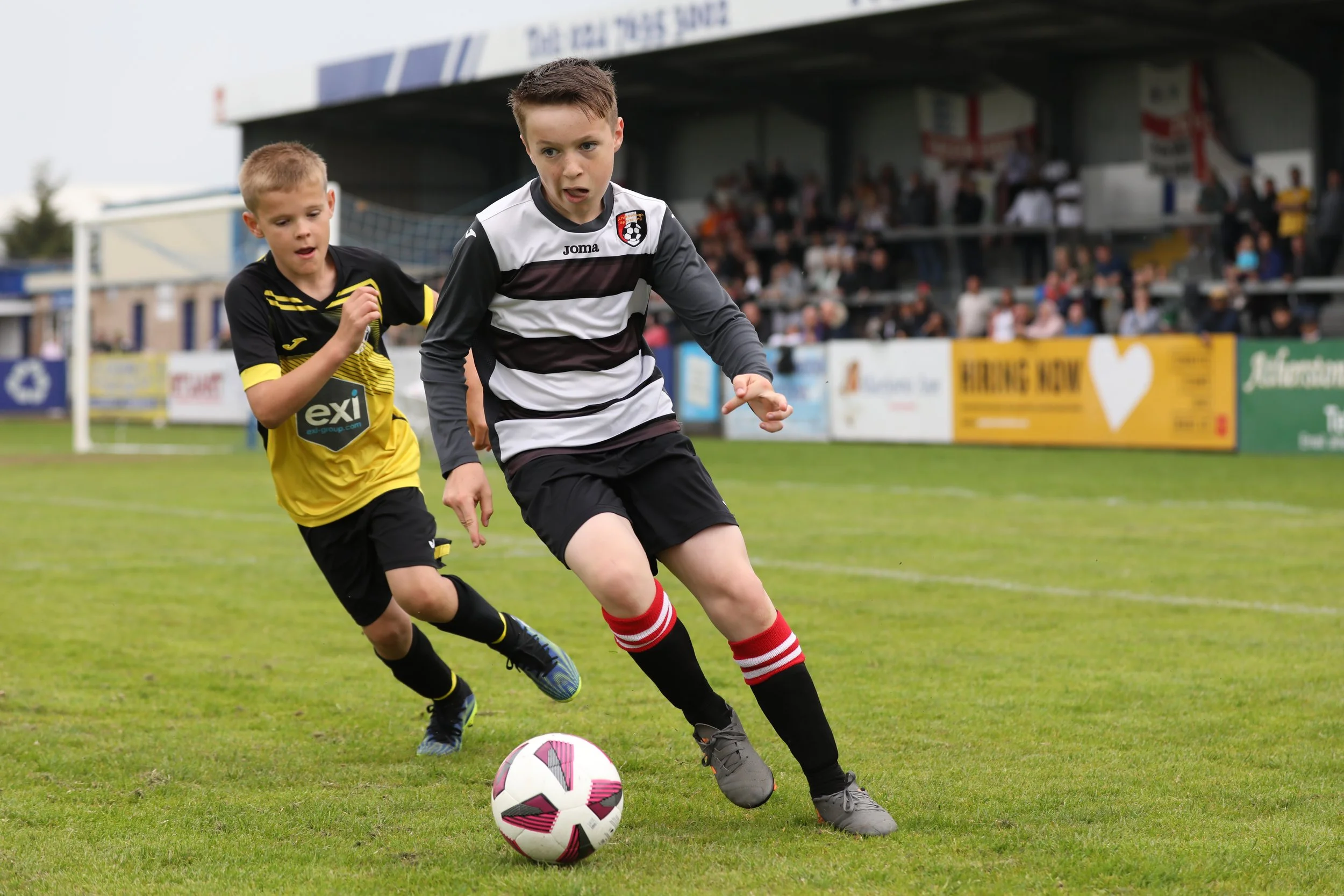 Two young boys playing soccer on a field, with one in a black and yellow jersey and the other in a black and white striped jersey, chasing a soccer ball.