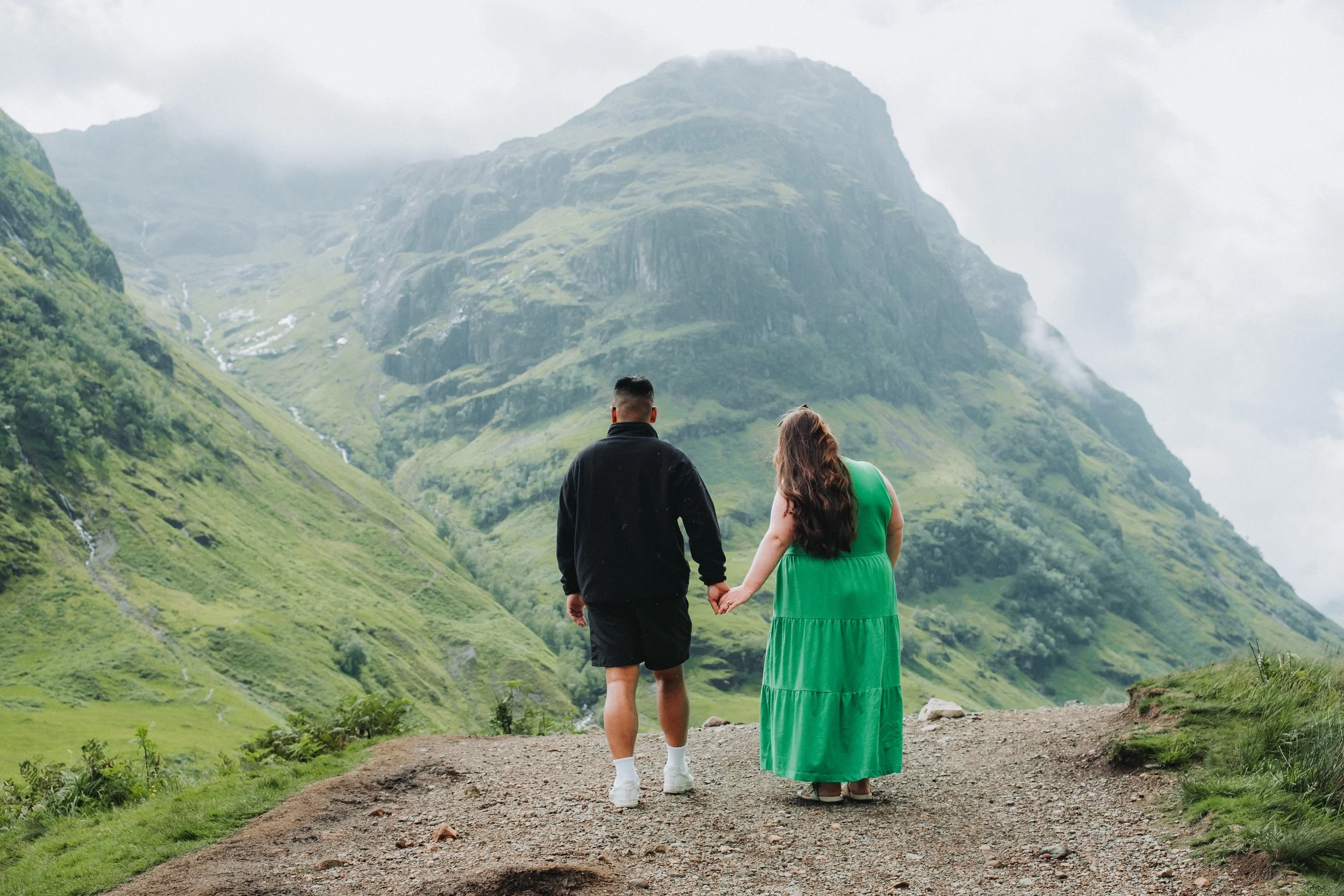A couple holding hands, walking on a dirt trail in a green mountainous landscape.