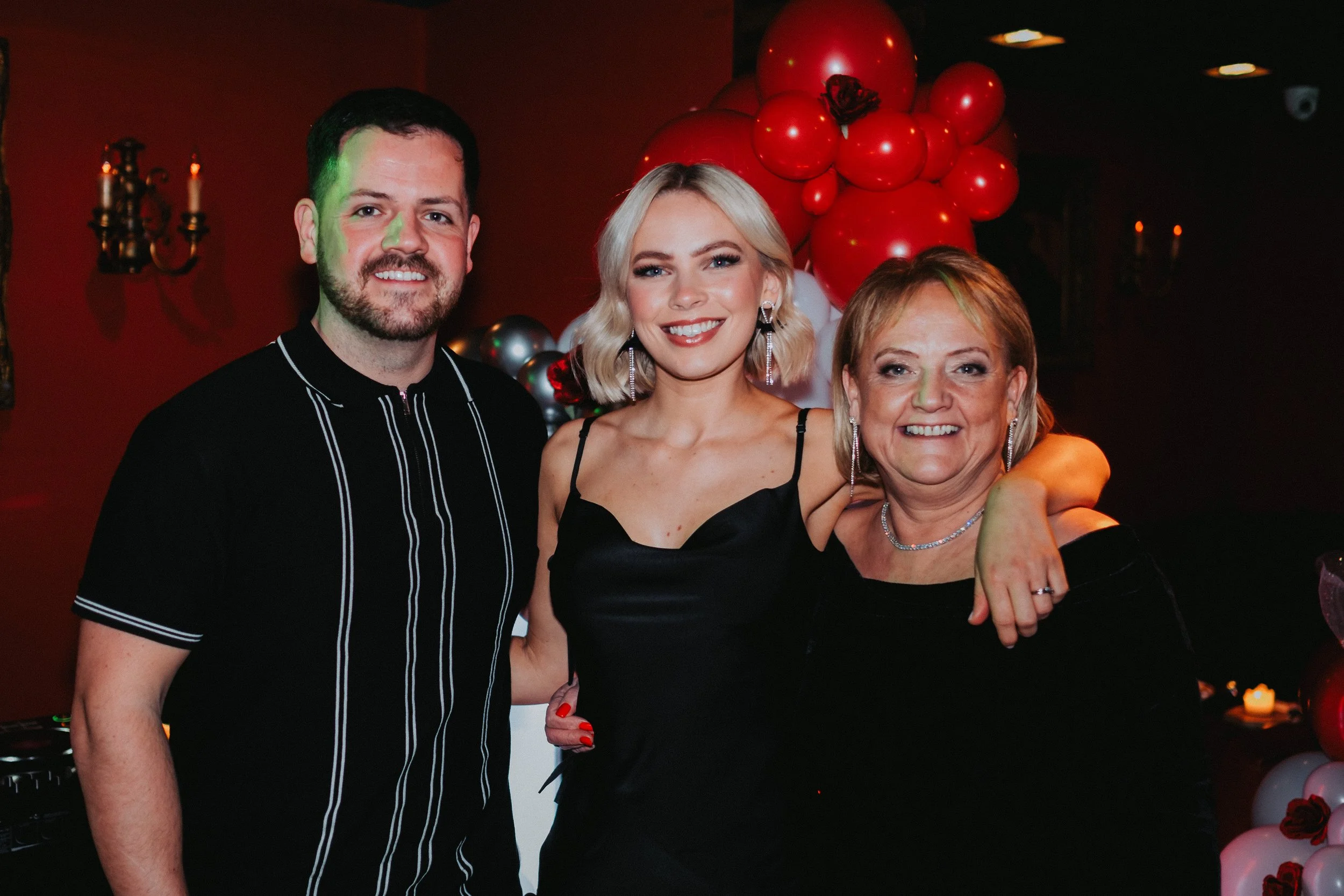 Three people smiling at a celebration with red, white, and black balloons in the background.