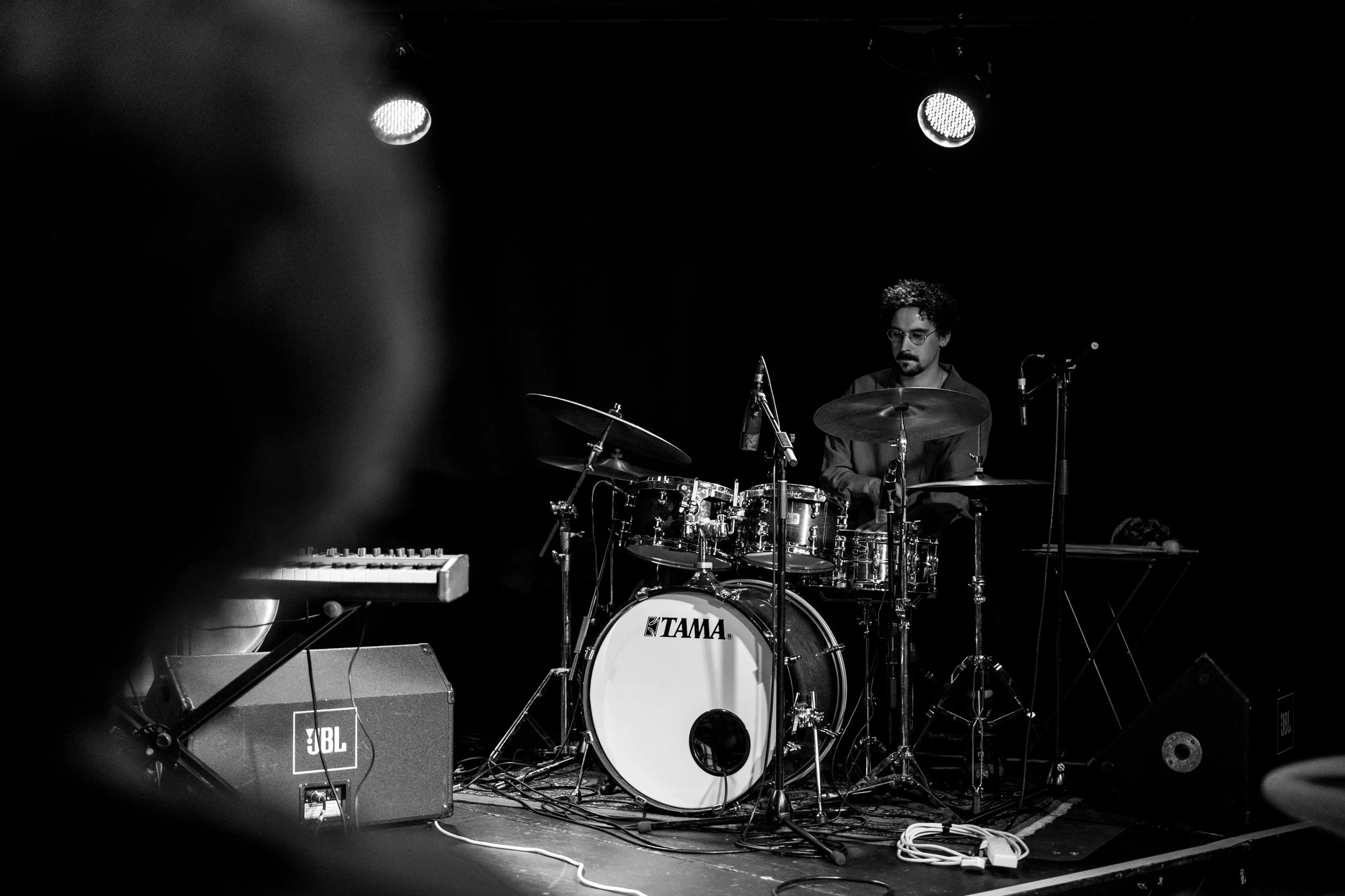 A man with glasses and curly hair playing a drum set on a stage with black background and stage lights.