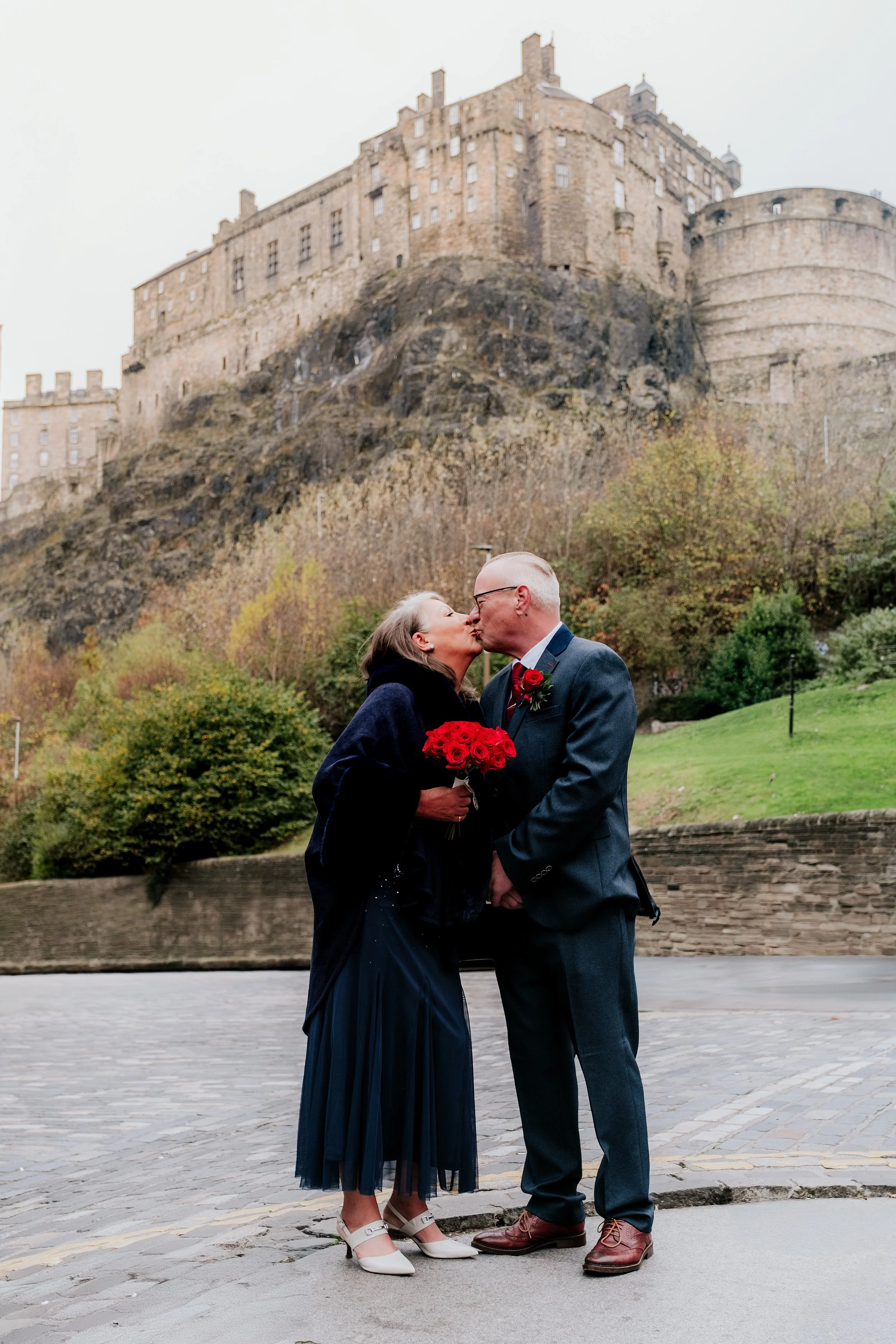 A couple kissing outdoors with a castle on a hill in the background. The woman is holding a bouquet of red roses, and both are dressed formally.