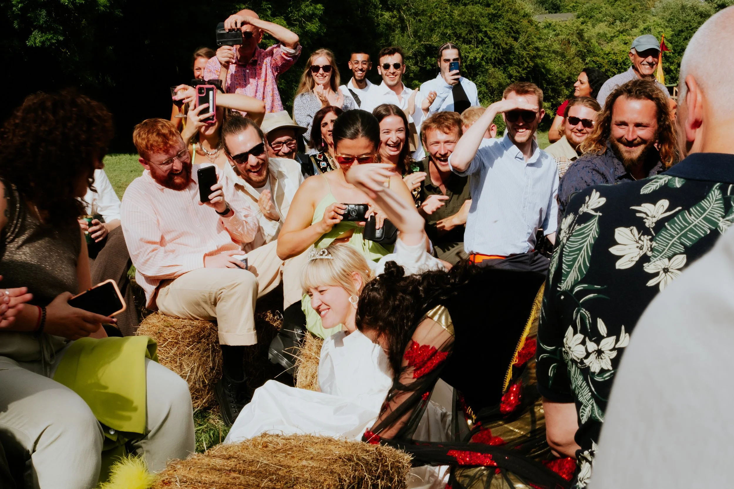 A group of people gathered outdoors smiling and taking pictures of a woman in wedding attire, sitting on hay bales, during a wedding celebration.