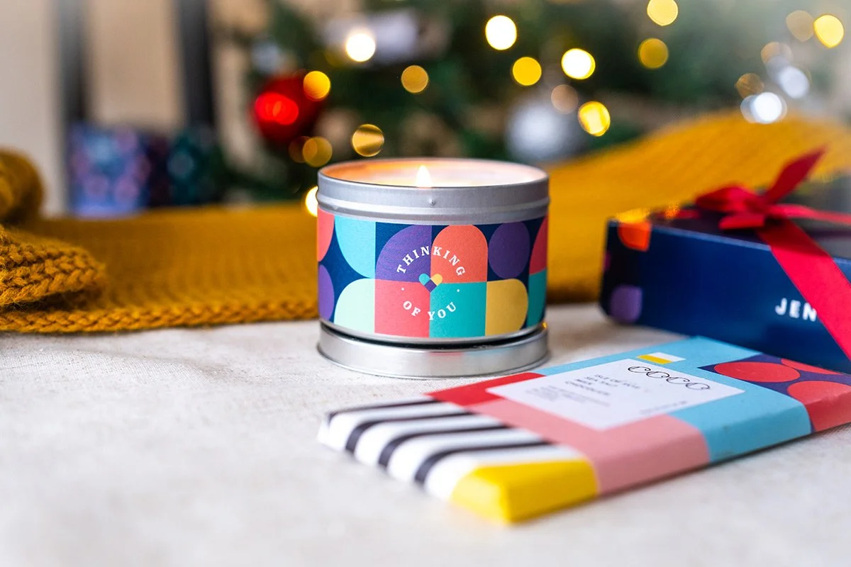A lit candle in a metal tin with colorful geometric patterns and the words "Thinking of You" on the label, placed on a white surface alongside a wrapped gift with a red ribbon and a colorful striped item, with a blurred background of Christmas tree l