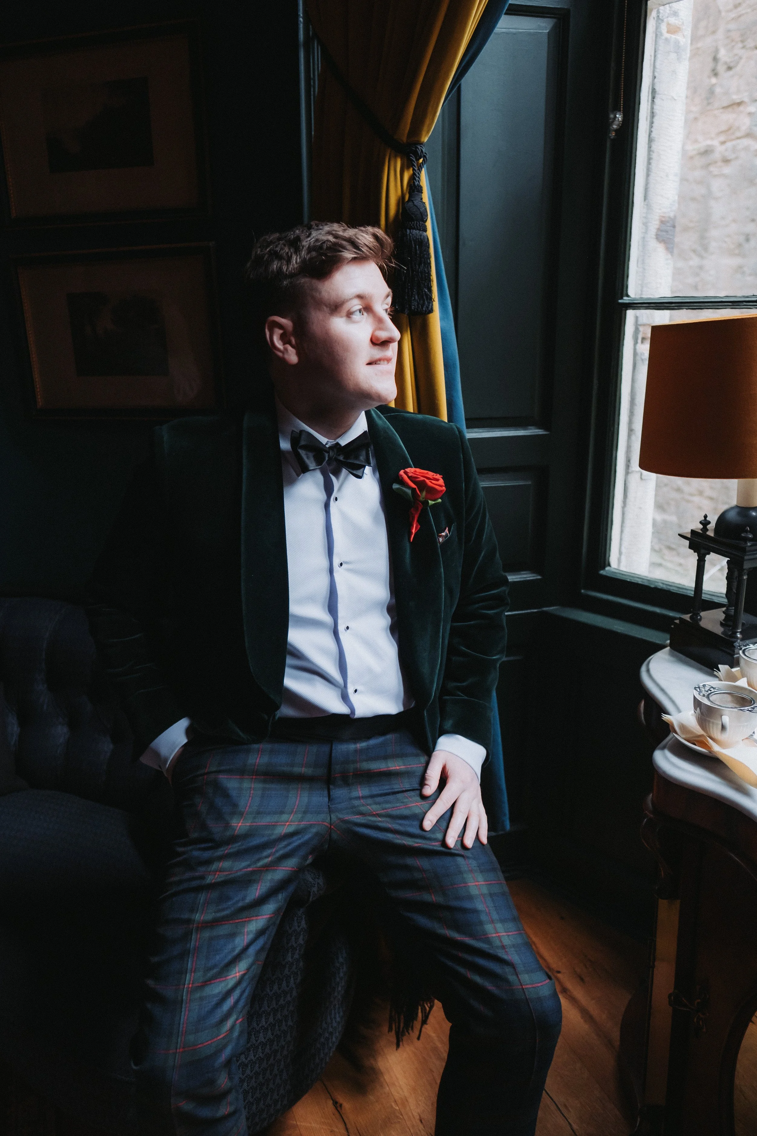 A young man in formal attire, including a tuxedo and plaid pants, sitting in a dimly lit room by a window with yellow curtains, looking contemplative. There is a table with tea cups and a lamp nearby.