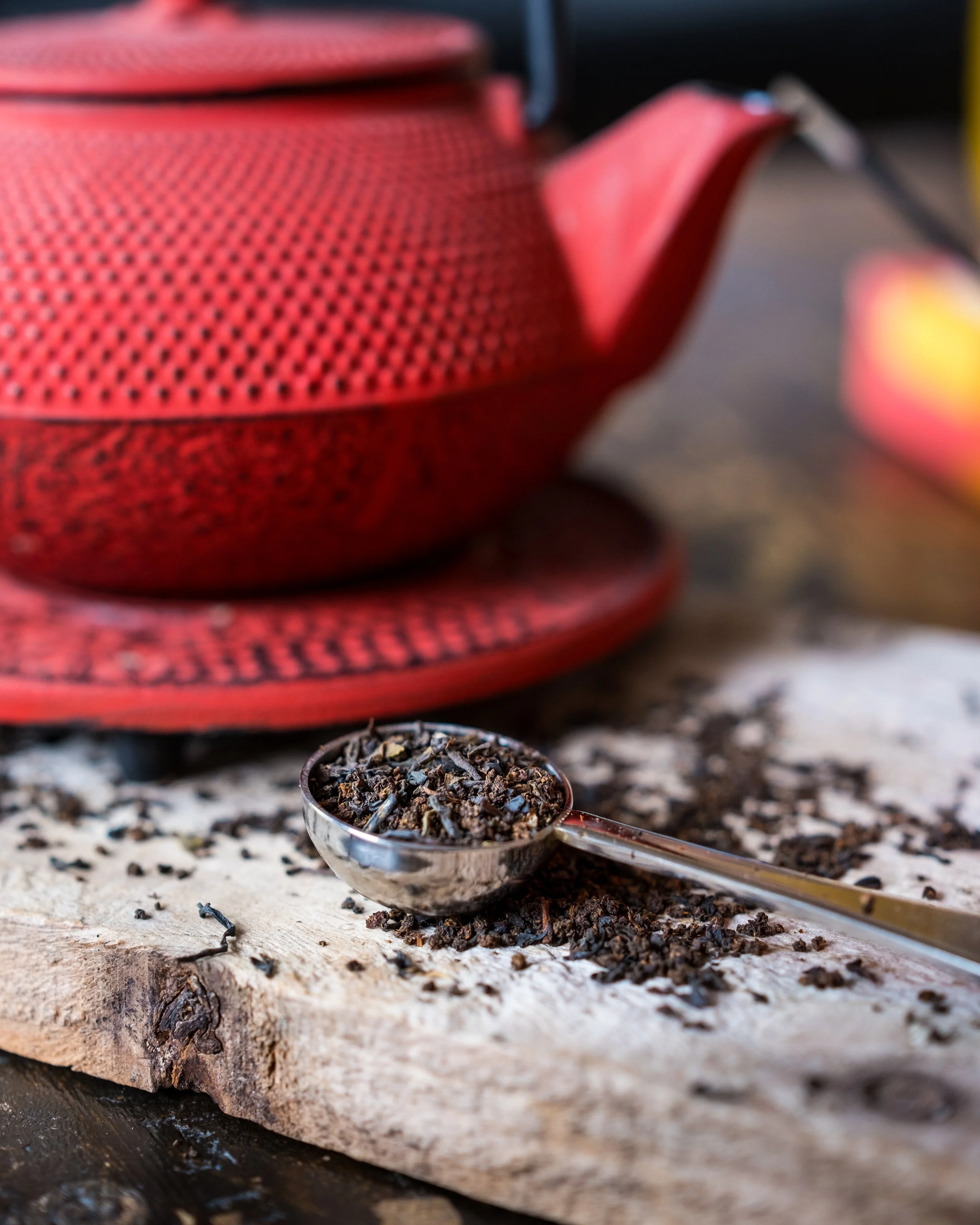 Close-up of a metal scoop filled with loose black tea leaves resting on a rustic wooden surface, with a red teapot in the background.