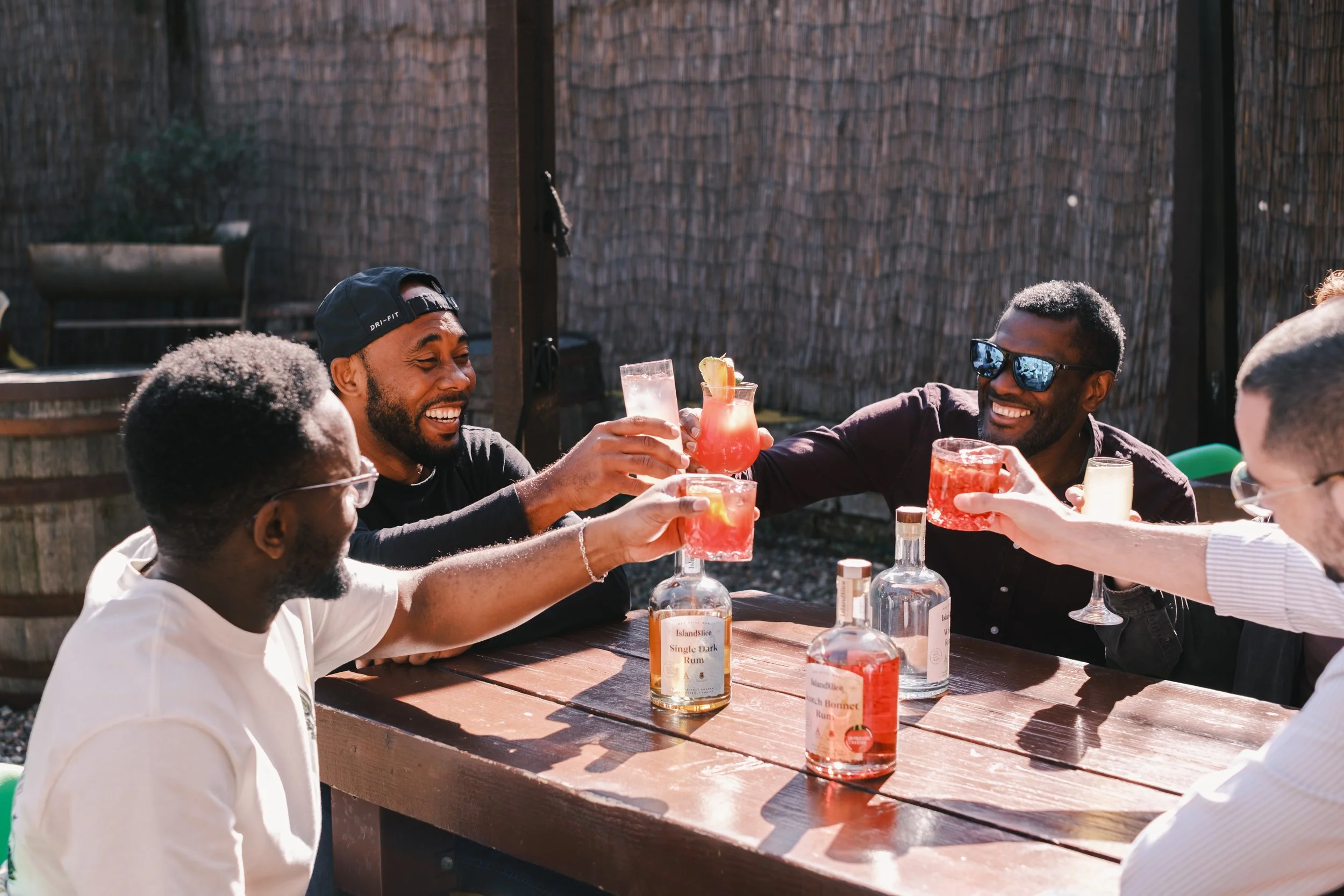 Group of five friends enjoying drinks and toasting outdoors at a wooden table with bottles and colorful cocktails, smiling and wearing casual clothes.