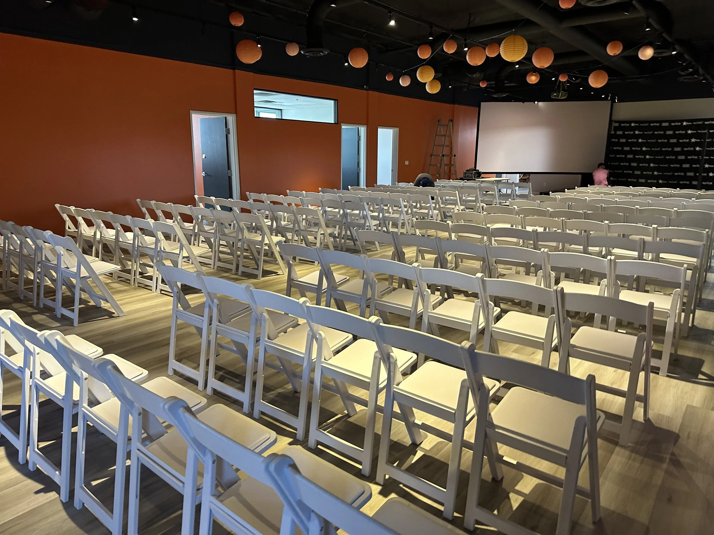Empty conference room with white folding chairs, an orange wall, hanging lanterns, a large projection screen, and a ladder.