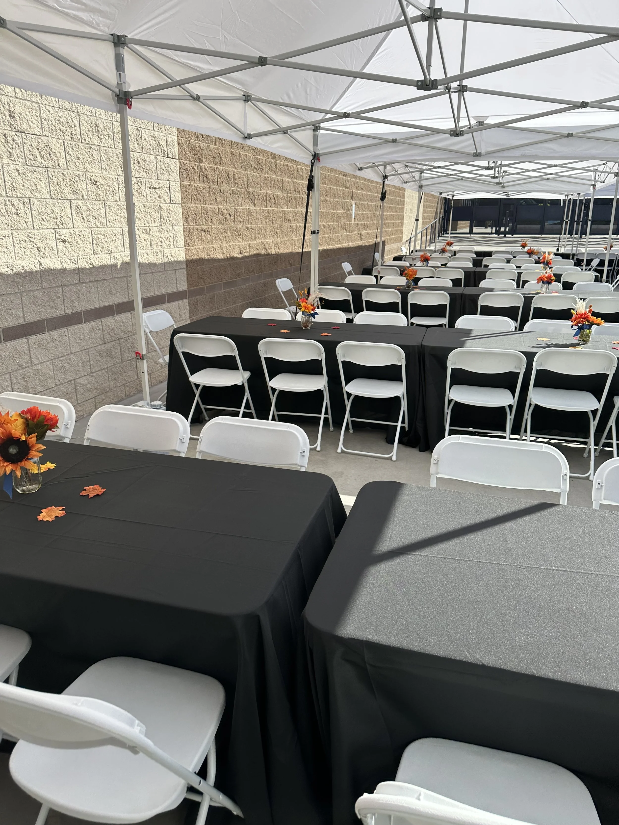 Outdoor event setup with black tablecloths, white chairs, and fall-themed floral centerpieces under a white canopy tent.